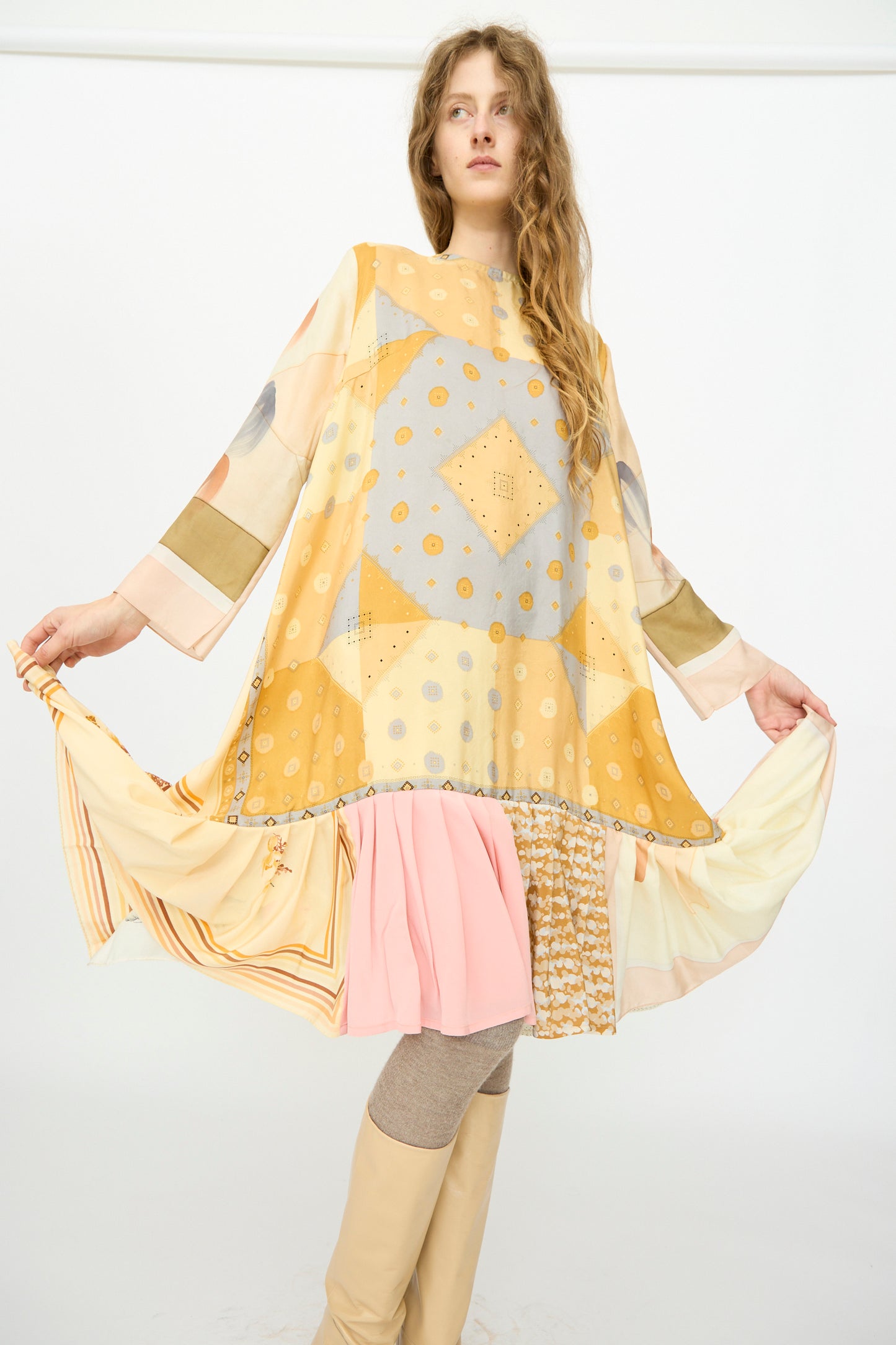 A woman models the Bettina Bakdal Hannah Silk Dress in Pius Pahl, featuring loose vintage style and geometric yellow, beige, and pink tones, paired with beige over-the-knee boots and tights against a plain white background.