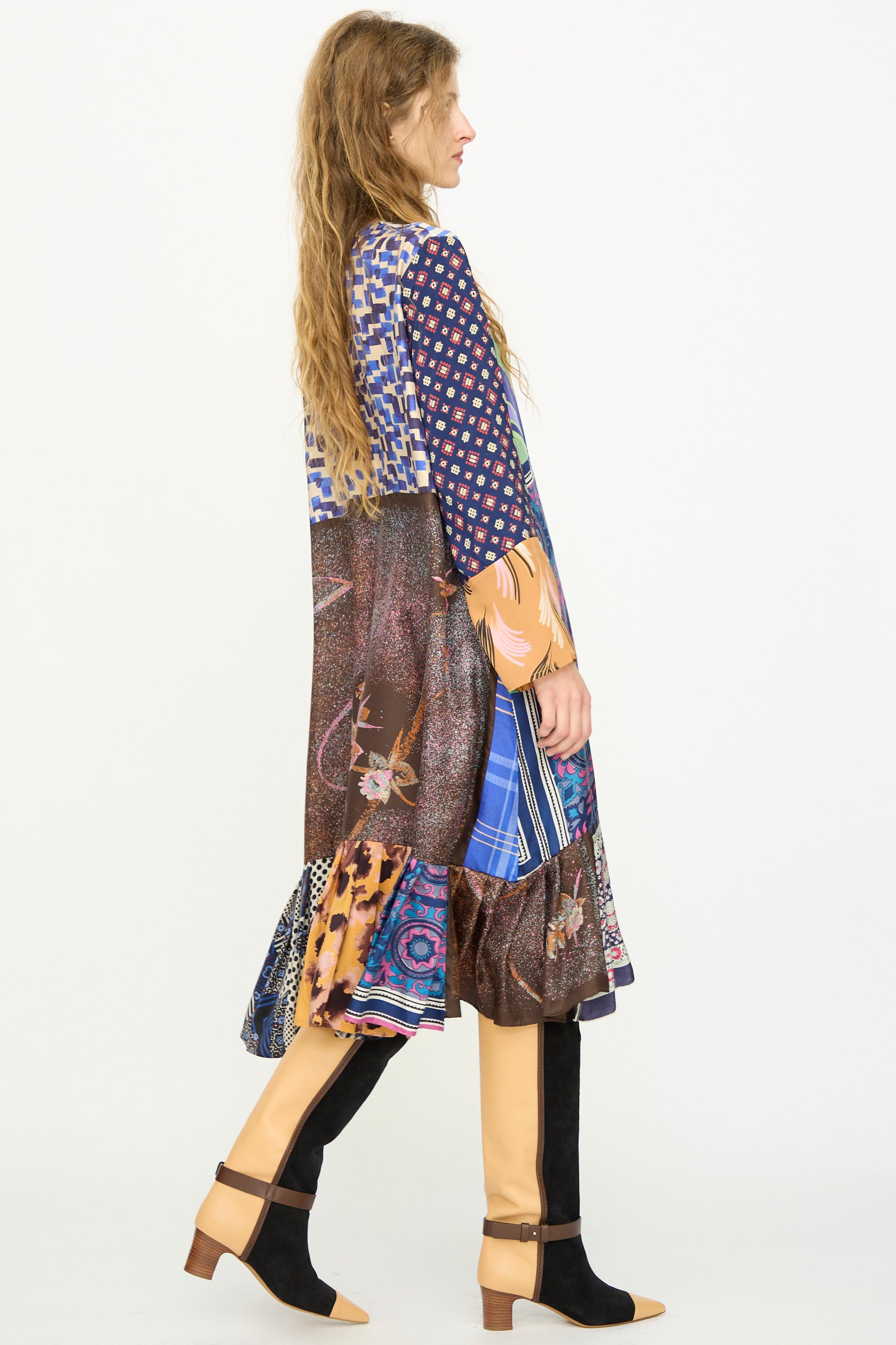 A woman with long wavy hair stands in profile against a plain white background wearing the Bettina Bakdal Hannah Silk Dress in Ricarda Meltzer and tan and black knee-high boots.