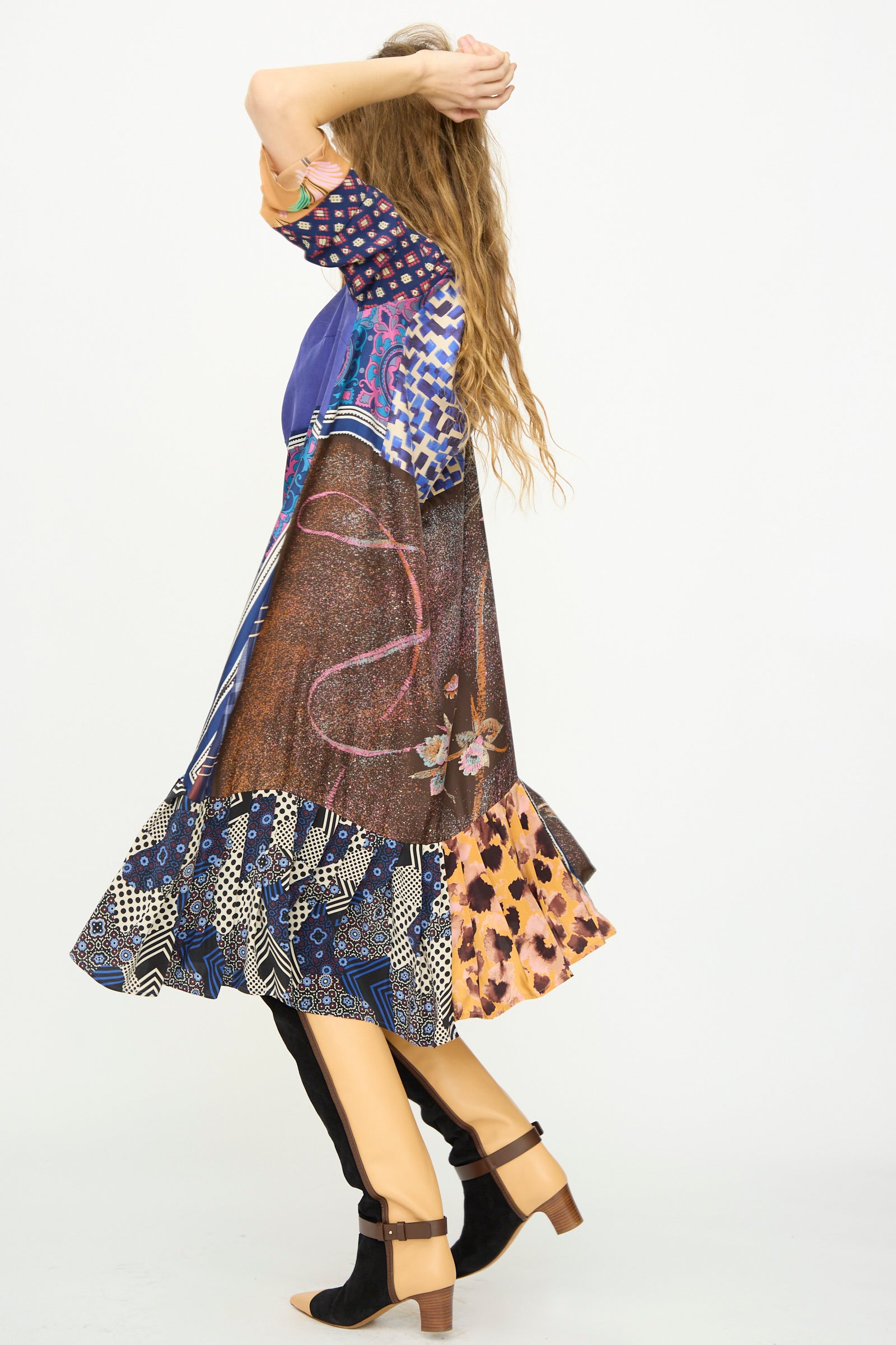 A woman with long wavy hair stands turned away, arms raised, wearing the Bettina Bakdal Hannah Silk Dress in Ricarda Meltzer and black and tan open-toe heels against a plain white background.