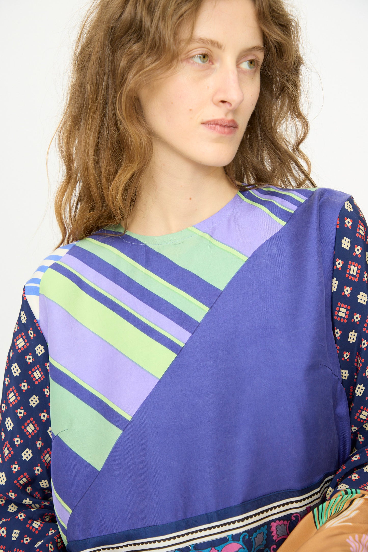 A person with wavy light brown hair wears the Bettina Bakdal Hannah Silk Dress in Ricarda Meltzer, featuring blue, green, and purple geometric patterns made from vintage silk scarves, looking slightly to the side.