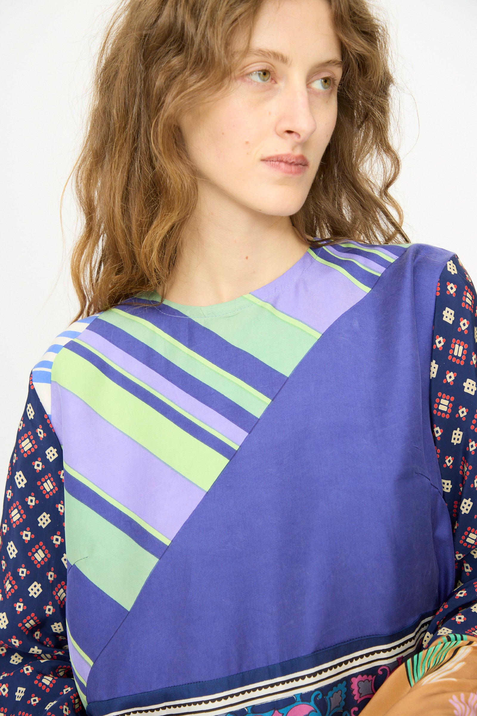 A person with wavy light brown hair wears the Bettina Bakdal Hannah Silk Dress in Ricarda Meltzer, featuring blue, green, and purple geometric patterns made from vintage silk scarves, looking slightly to the side.