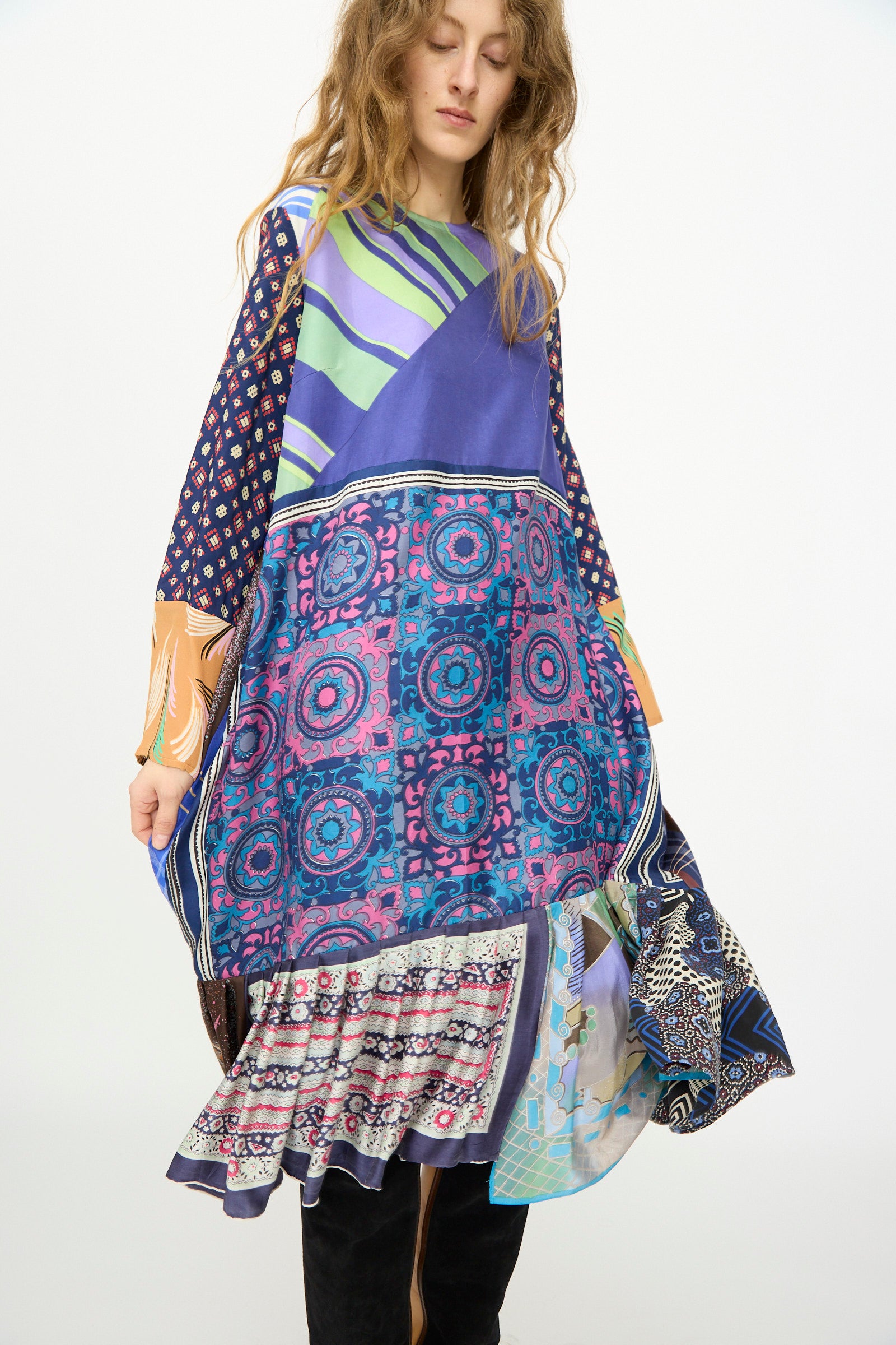 A woman stands against a plain background wearing the Hannah Silk Dress in Ricarda Meltzer by Bettina Bakdal, crafted from upcycled vintage silk scarves in colorful geometric patchwork.