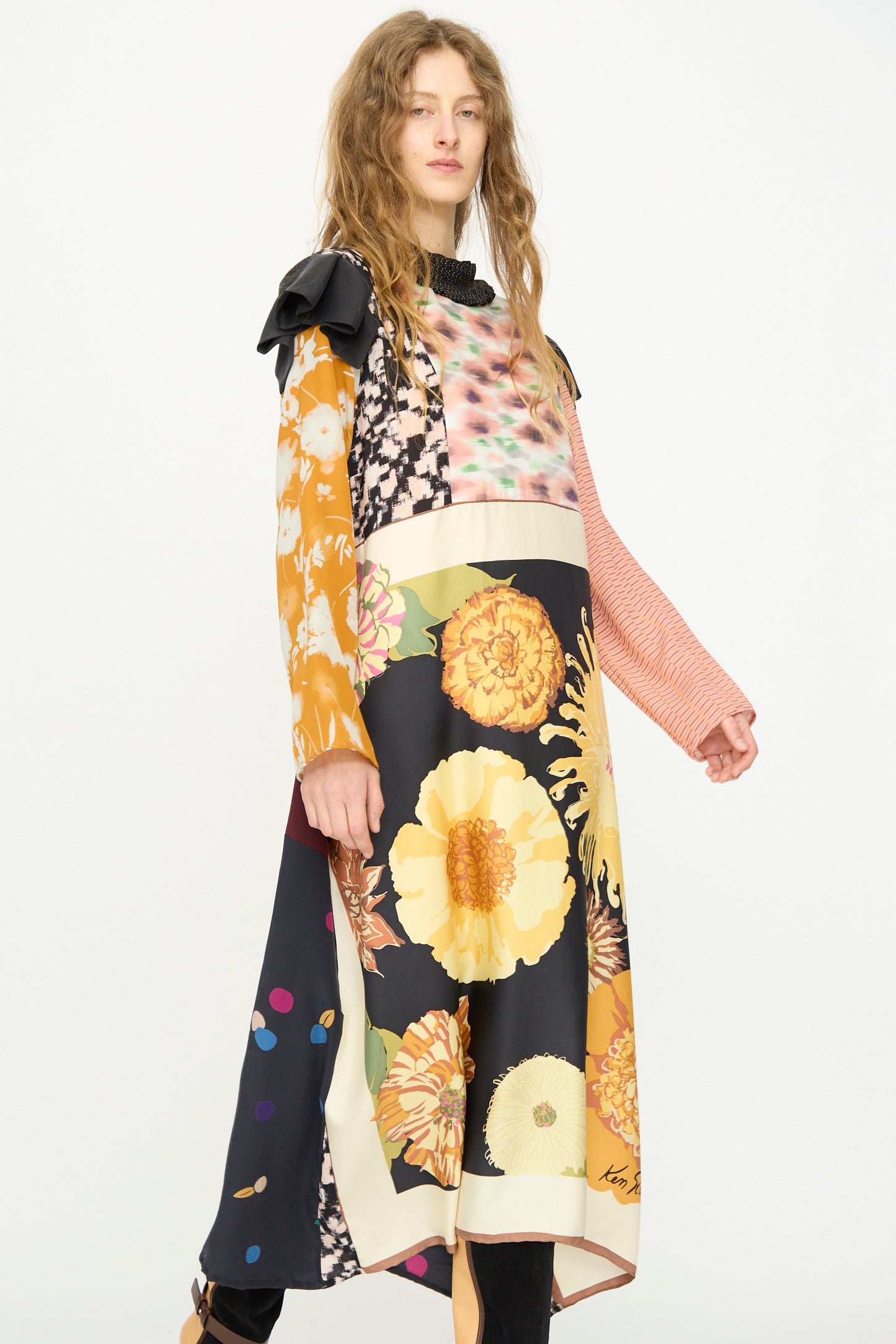 A person in the Bettina Bakdal Ruffled Collar and Shoulder Vintage Scarves Dress in Edith Suschitzky, featuring up-cycled silk with long sleeves and mixed floral and geometric prints, stands against a plain white background.