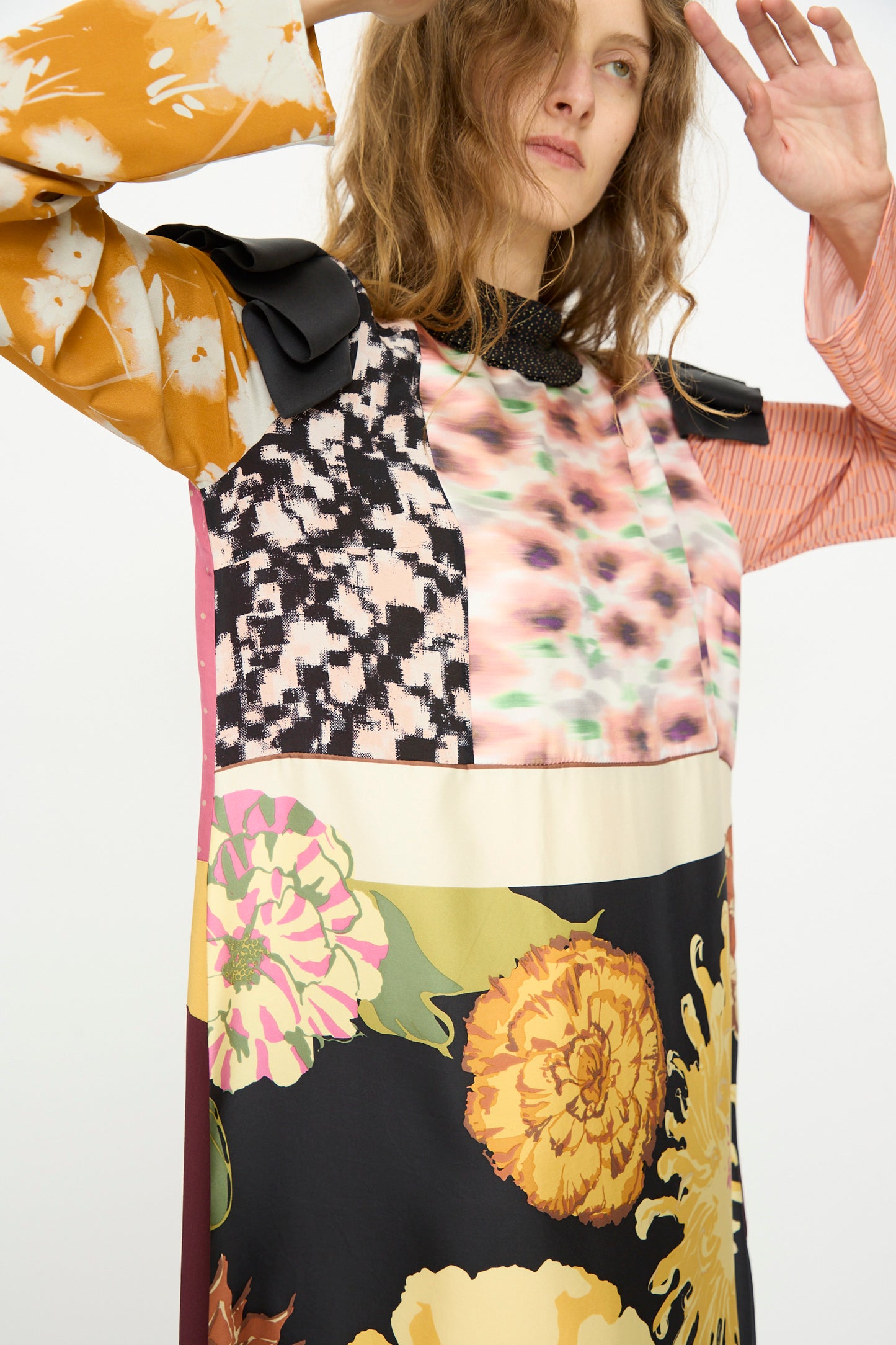 A woman in the Bettina Bakdal Ruffled Collar and Shoulder Vintage Scarves Dress in Edith Suschitzky, featuring patchwork prints, stands with arms raised against a plain white background.