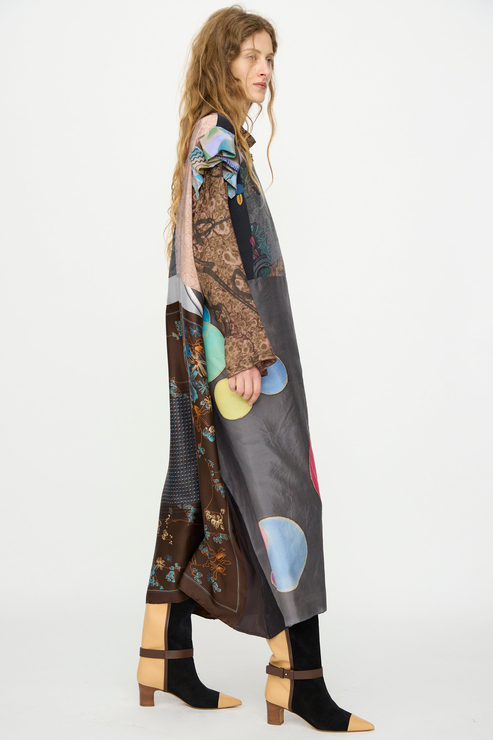 A woman with long wavy hair models the Bettina Bakdal Ruffled Collar and Shoulder Vintage Scarves Dress in Margarete Dambeck, featuring vibrant patterns, paired with black-and-tan ankle-strap shoes against a white background.