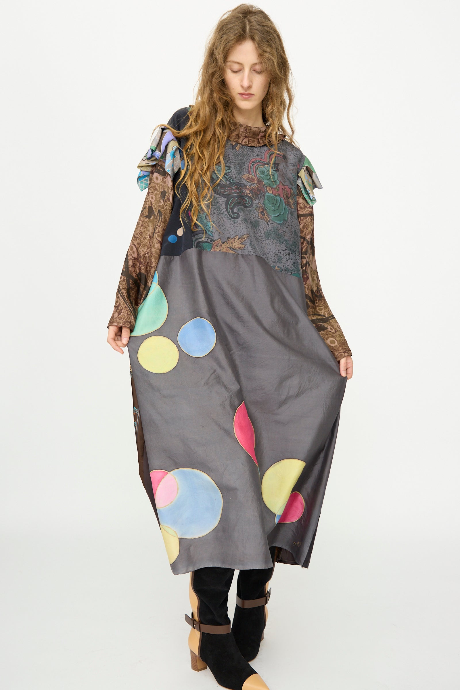 A woman wears the Bettina Bakdal Ruffled Collar and Shoulder Vintage Scarves Dress in Margarete Dambeck, featuring a long, loose fit with colorful circular patterns and layered sleeves as she holds out the skirt.