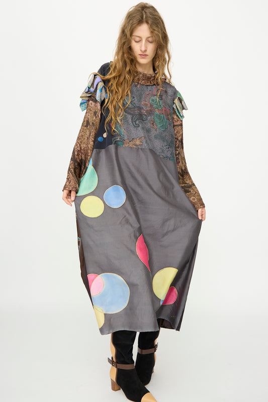 A woman wears the Bettina Bakdal Ruffled Collar and Shoulder Vintage Scarves Dress in Margarete Dambeck, featuring a long, loose fit with colorful circular patterns and layered sleeves as she holds out the skirt.