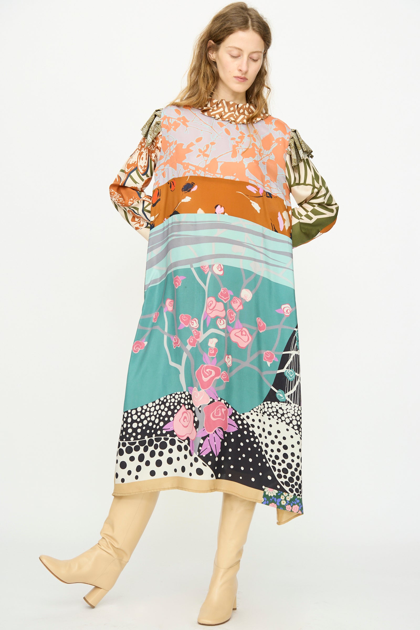 A woman wears the Bettina Bakdal Ruffled Collar and Shoulder Vintage Scarves Dress in Dolores Del Bochum, featuring a colorful, abstract floral patchwork, paired with knee-high beige boots as she stands with her head down and arms at her sides.