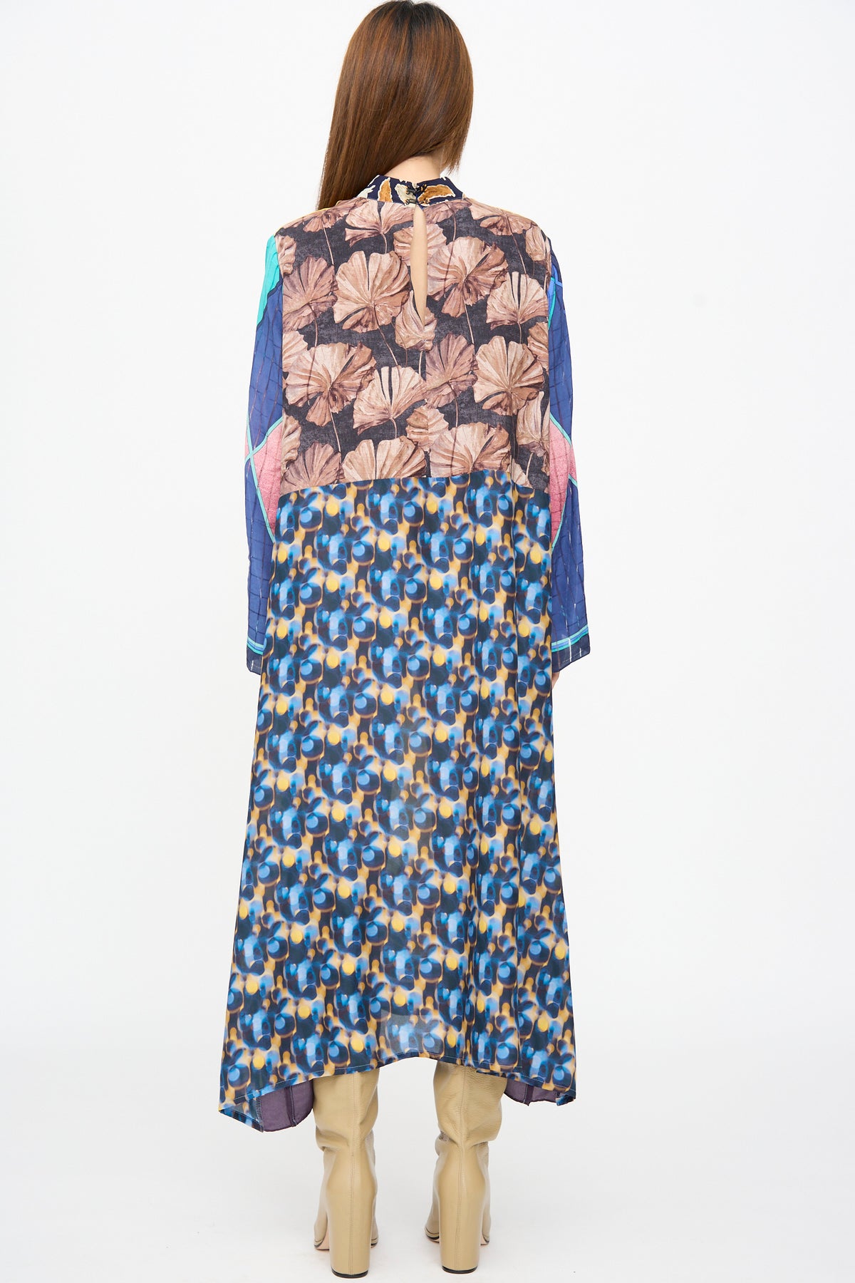A person seen from the back wearing a Bettina Bakdal Classic Mandarin Collar Dress in Alice Vollmer, featuring brown palm leaf and abstract patterned panels, paired with beige block-heel boots.