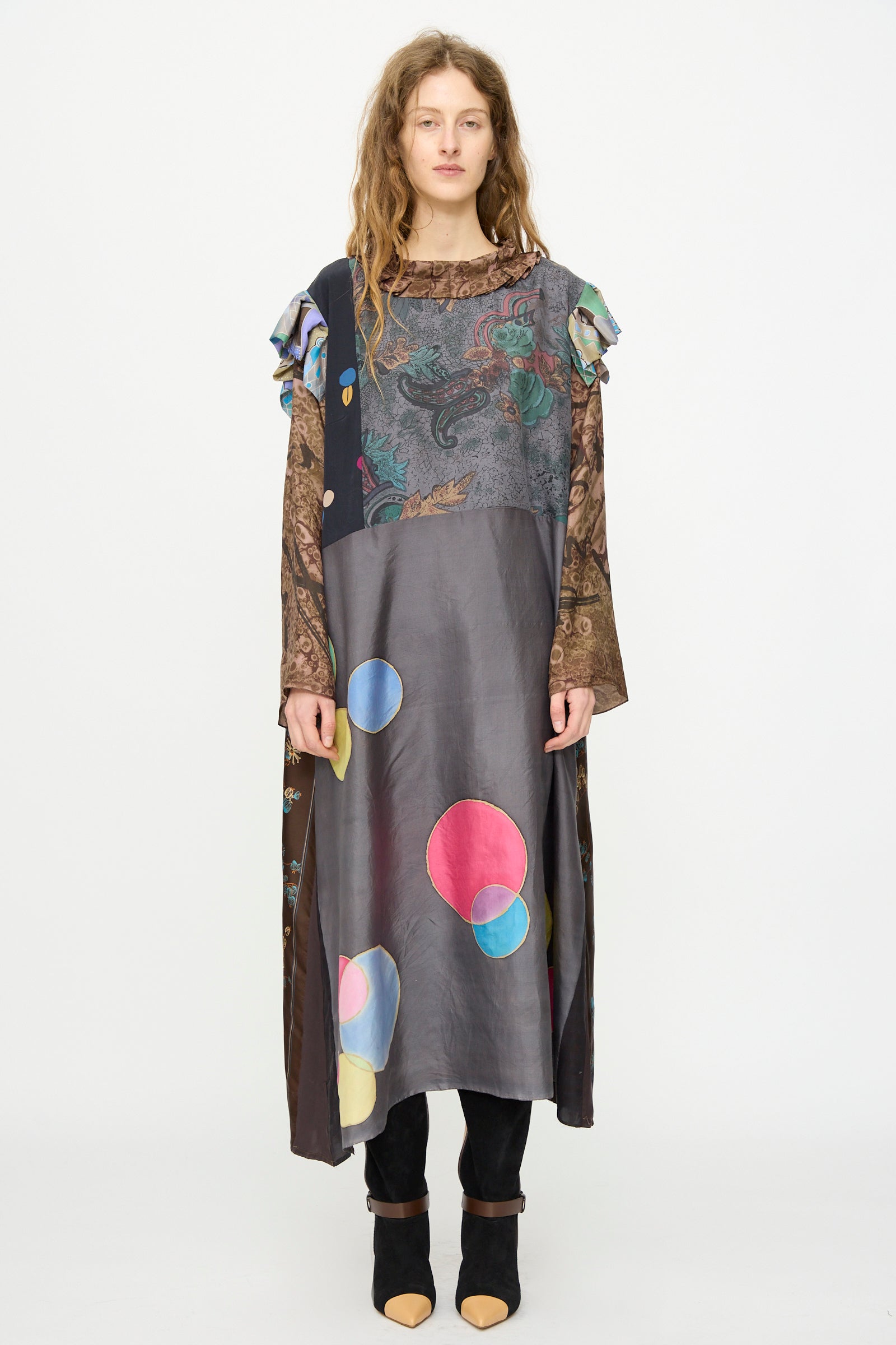 A woman faces forward in the Bettina Bakdal Ruffled Collar and Shoulder Vintage Scarves Dress in Margarete Dambeck, featuring mixed patterns over a long-sleeve top and black pants.