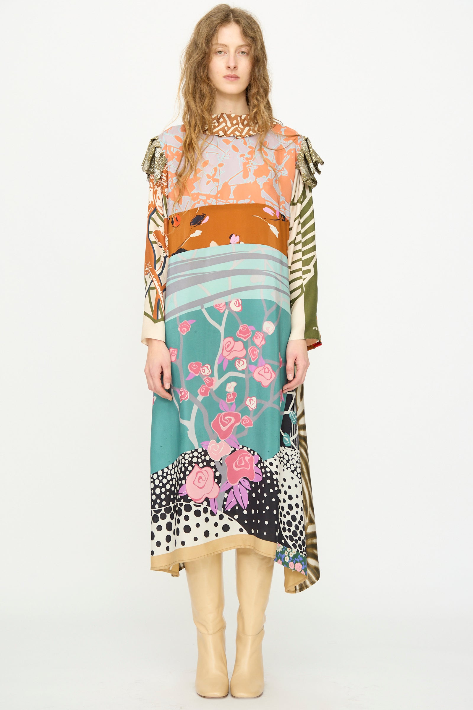 Person standing against a plain white background wearing the Bettina Bakdal Ruffled Collar and Shoulder Vintage Scarves Dress in Dolores Del Bochum, styled with cream boots and long, wavy hair.