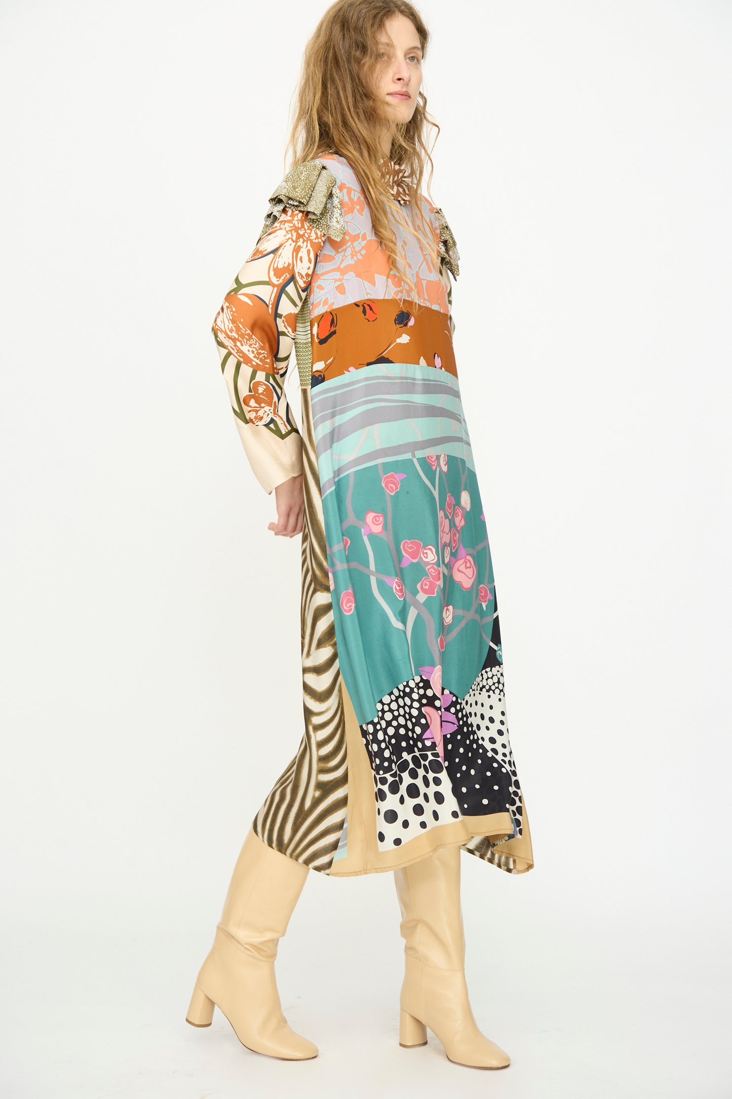 A woman wears the Bettina Bakdal Ruffled Collar and Shoulder Vintage Scarves Dress in Dolores Del Bochum with beige knee-high boots, standing against a plain white background.