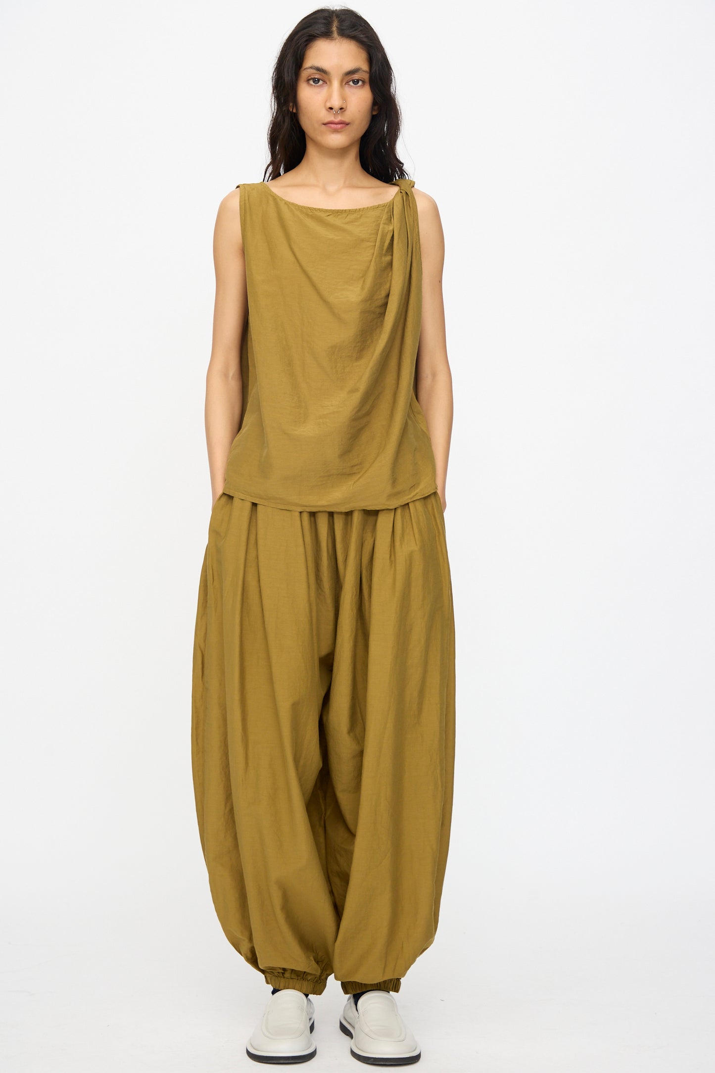 A person wears relaxed fit Akari Pants in Dark Olive by Black Crane, paired with a sleeveless draped mustard top and white slip-on shoes, standing against a plain white background.