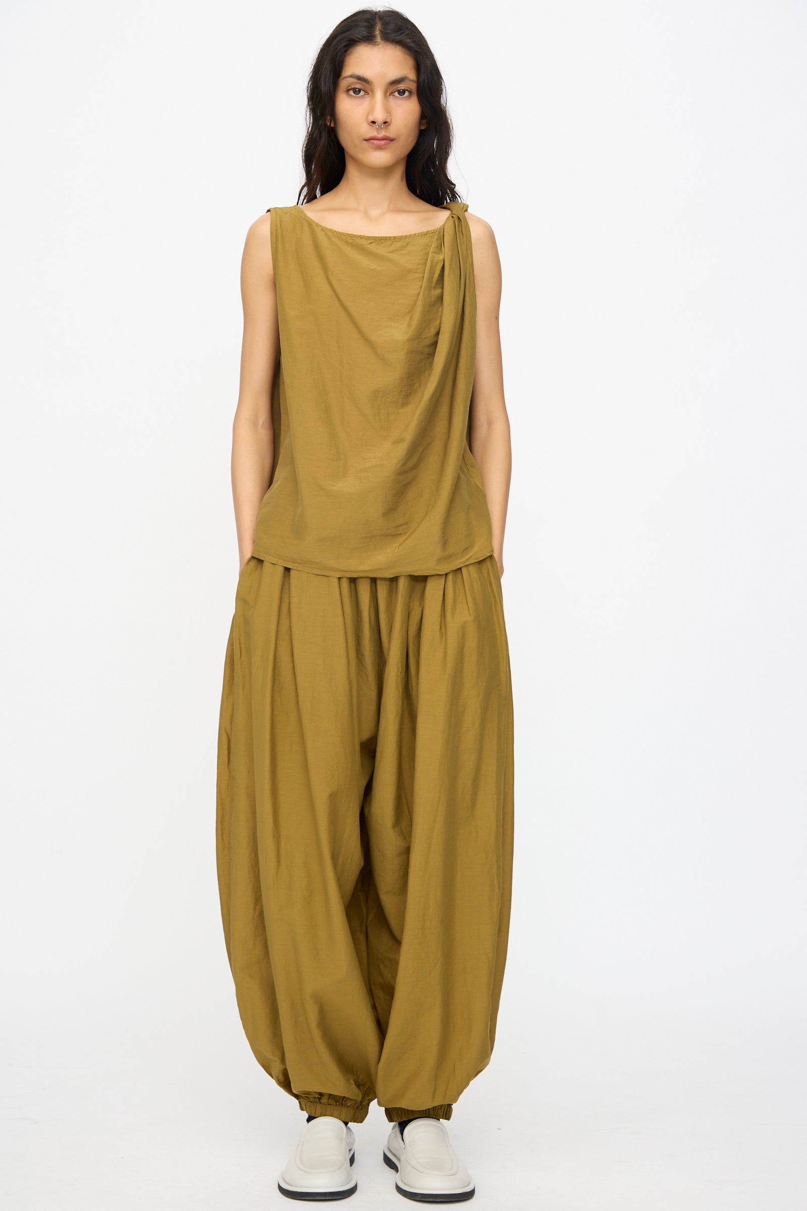 A person wears relaxed fit Akari Pants in Dark Olive by Black Crane, paired with a sleeveless draped mustard top and white slip-on shoes, standing against a plain white background.