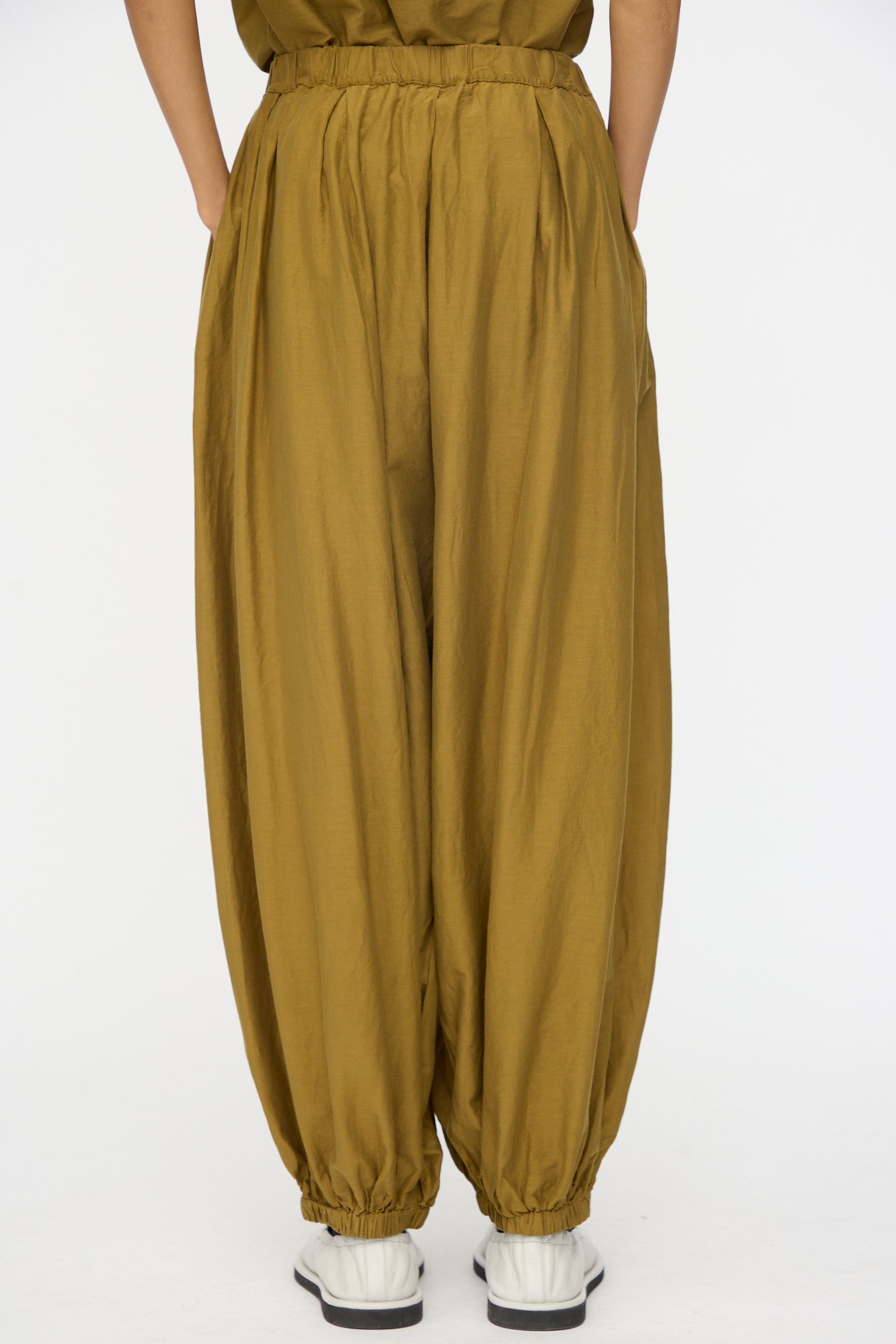 A person wears Black Crane's Akari Pants in Dark Olive, featuring a relaxed fit and elastic cuffs, paired with white sneakers against a plain white background.