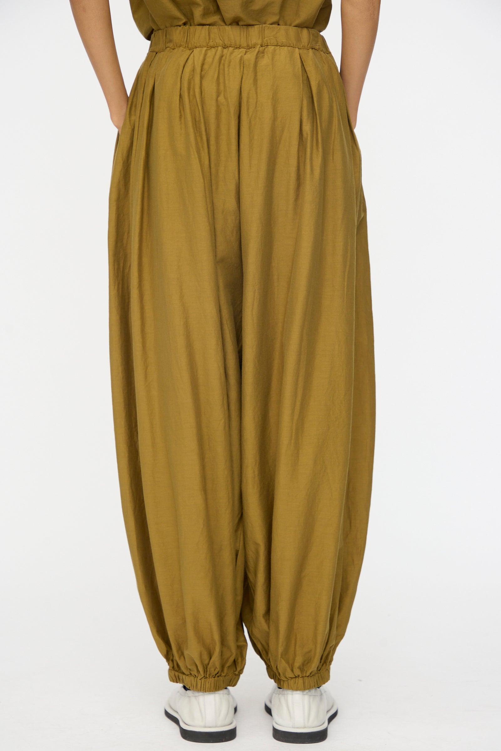 A person wears Black Crane's Akari Pants in Dark Olive, featuring a relaxed fit and elastic cuffs, paired with white sneakers against a plain white background.