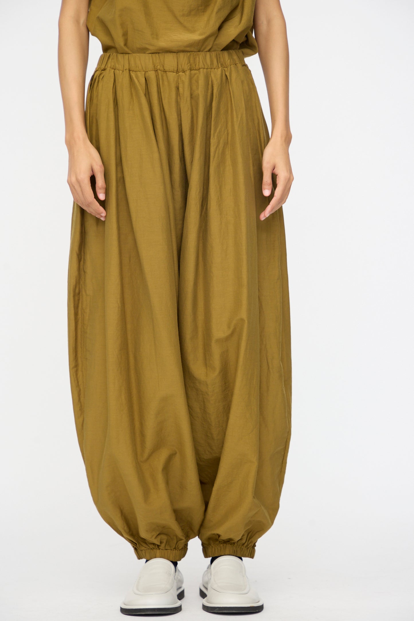 A person wears Black Crane's Akari Pants in Dark Olive, featuring a relaxed fit and elastic ankles, crafted from a soft cotton silk blend, paired with white slip-on shoes against a plain white background.