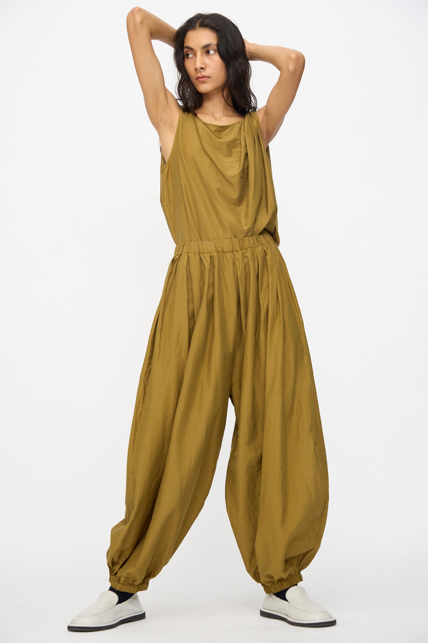 A person wears Black Crane's Akari Pants in Dark Olive—a relaxed, balloon-style silhouette in a soft cotton-silk blend—paired with white slip-on shoes, standing with arms raised behind head against a plain white background.