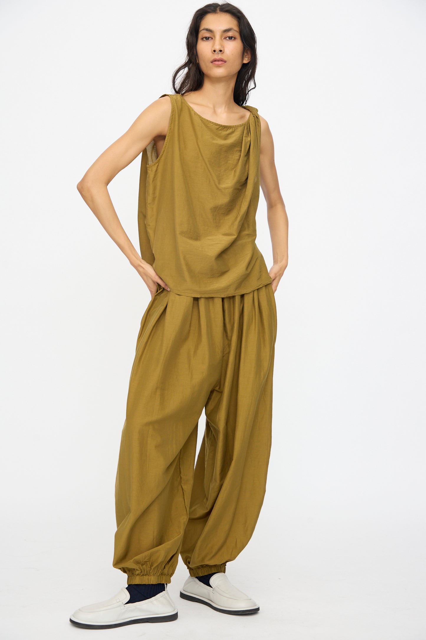 A person stands against a plain white background wearing Black Crane's Akari Pants in Dark Olive, styled with a loose, sleeveless top and white slip-on shoes.