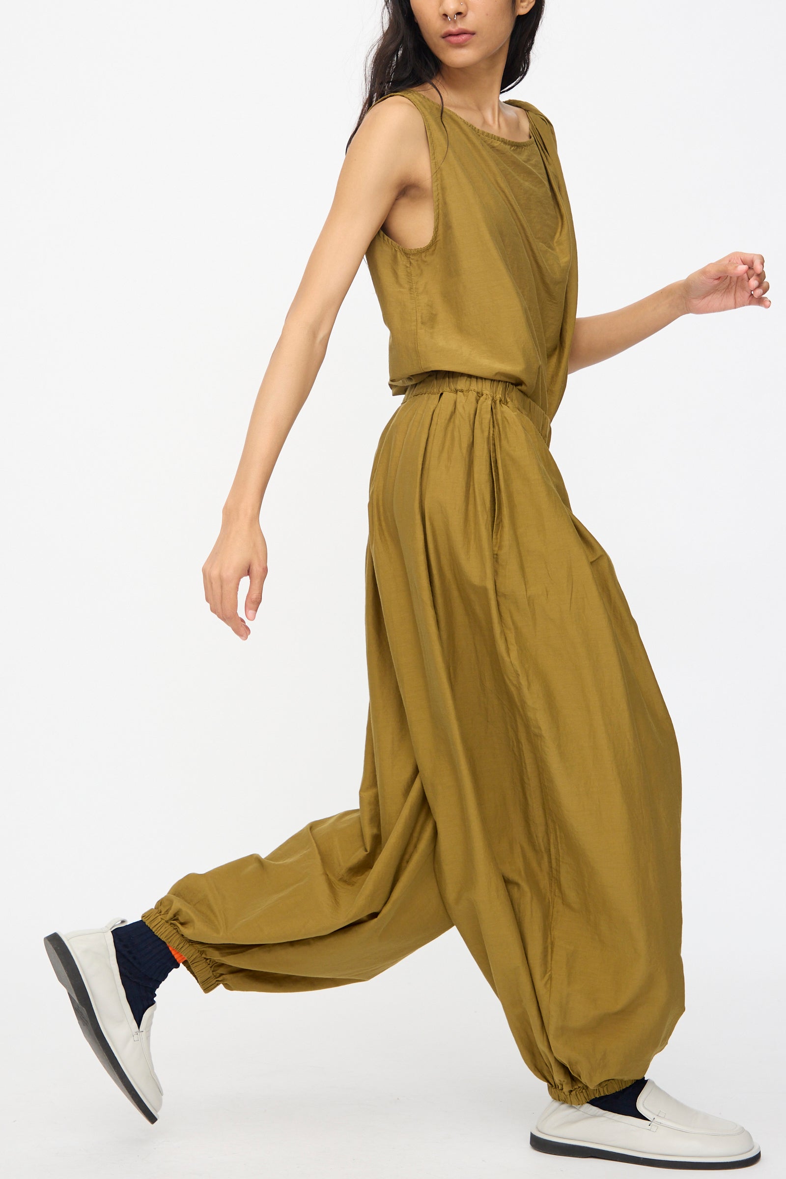 A person walks forward wearing Black Crane's Akari Pants in Dark Olive—relaxed fit, wide-leg with elastic cuffs—styled with a matching sleeveless top, white shoes, and dark socks.