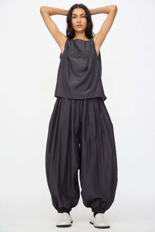 A person stands with arms raised, wearing a loose, dark sleeveless top and Black Crane Akari Pants in Faded Black—a cotton silk blend balloon pant with elastic waist and cuffs—paired with light shoes against a white background.