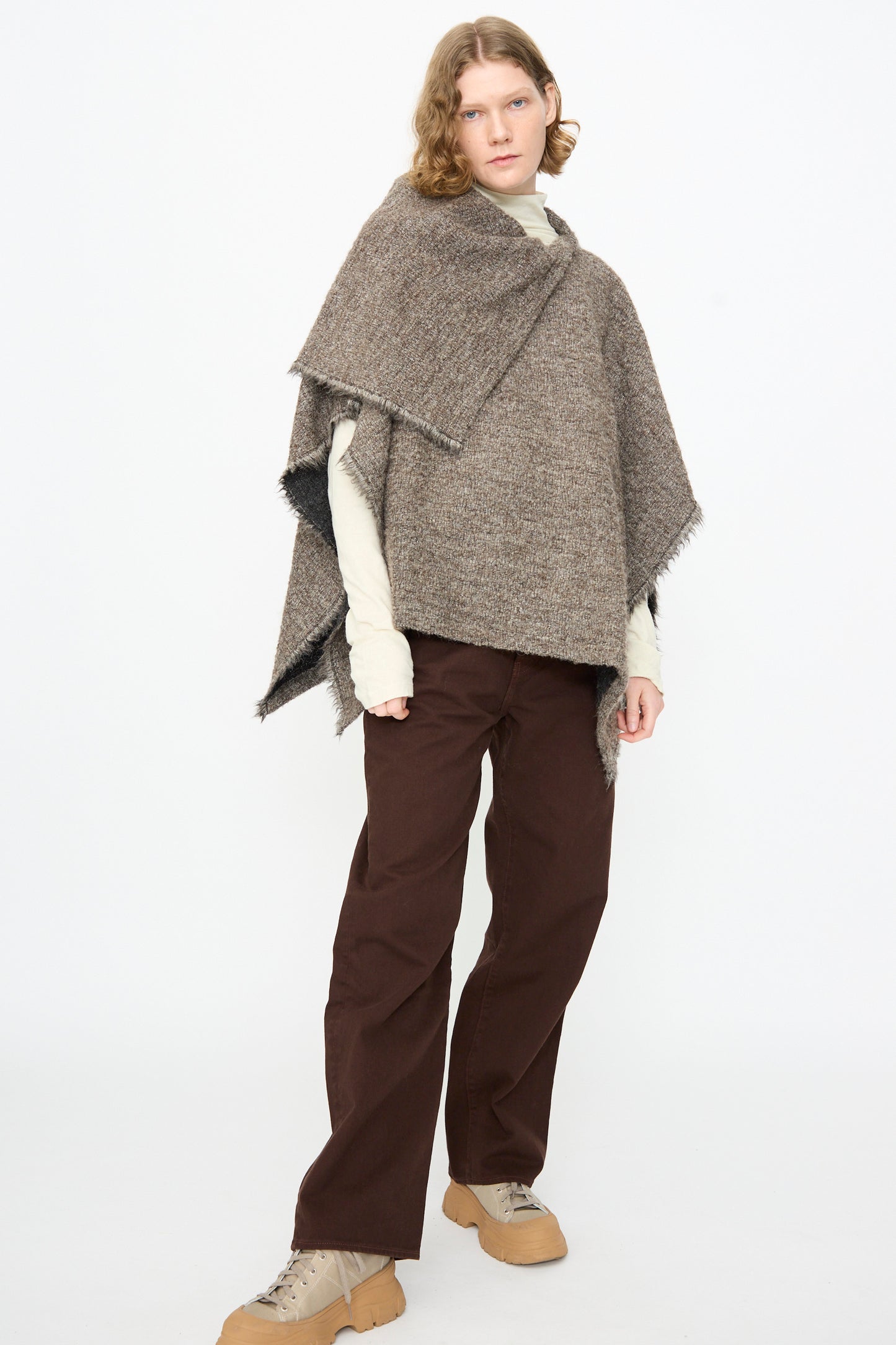 A person stands against a white background, wearing a beige turtleneck, the Black Crane Alpaca Short Poncho in Beige (made in USA) with fringe details, brown pants, and tan shoes.
