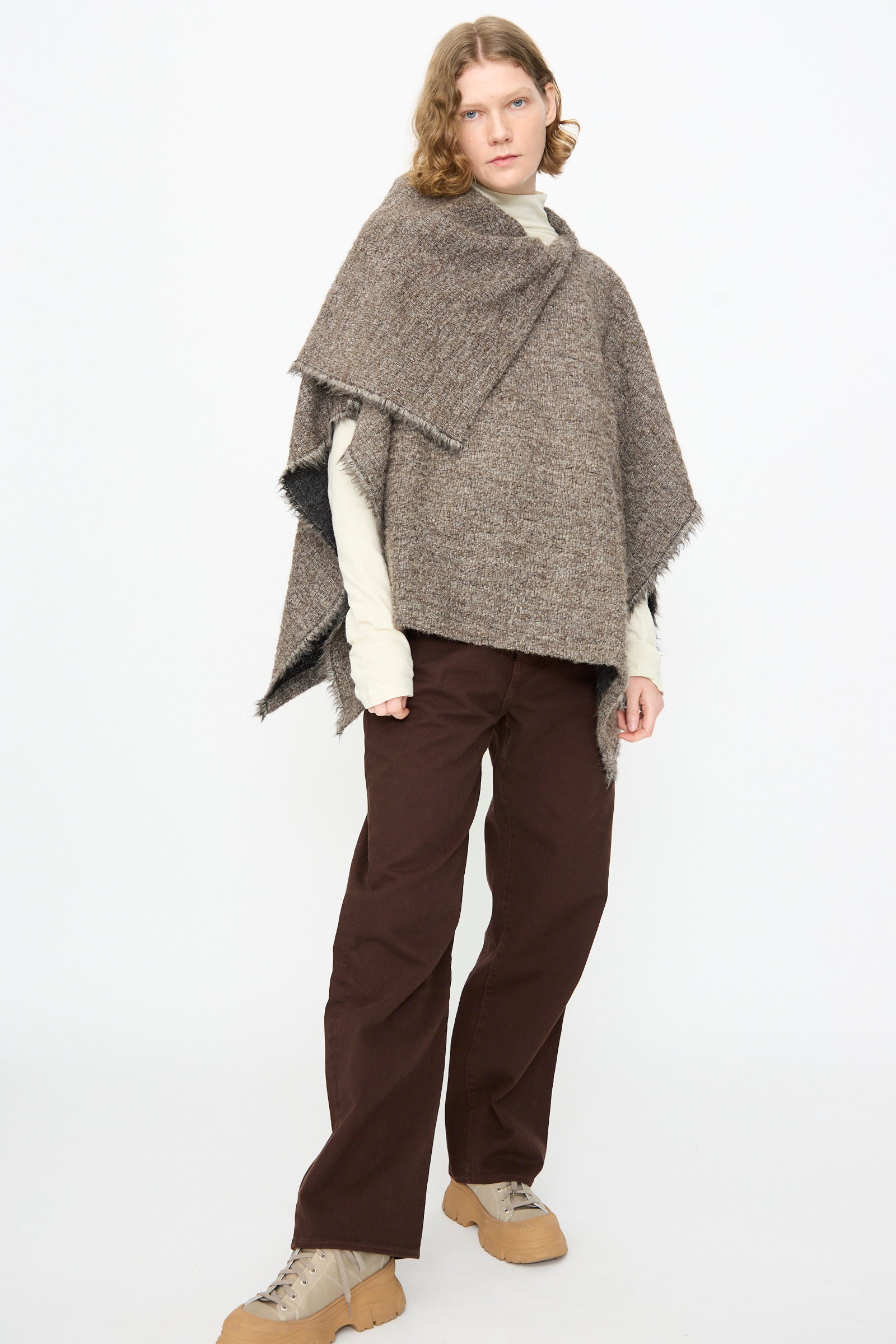 A person stands against a white background, wearing a beige turtleneck, the Black Crane Alpaca Short Poncho in Beige (made in USA) with fringe details, brown pants, and tan shoes.