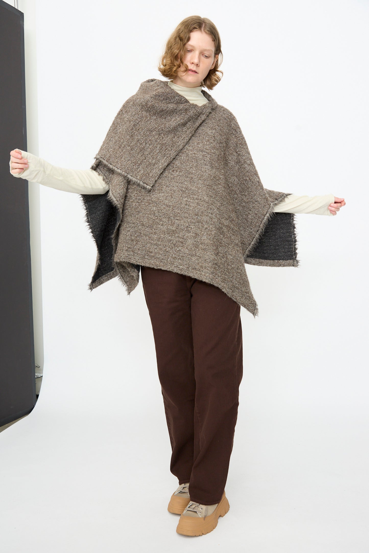 A person stands against a plain white background with arms slightly outstretched, wearing the Black Crane Alpaca Short Poncho in Beige over a cream long-sleeve shirt, brown pants, and tan shoes.