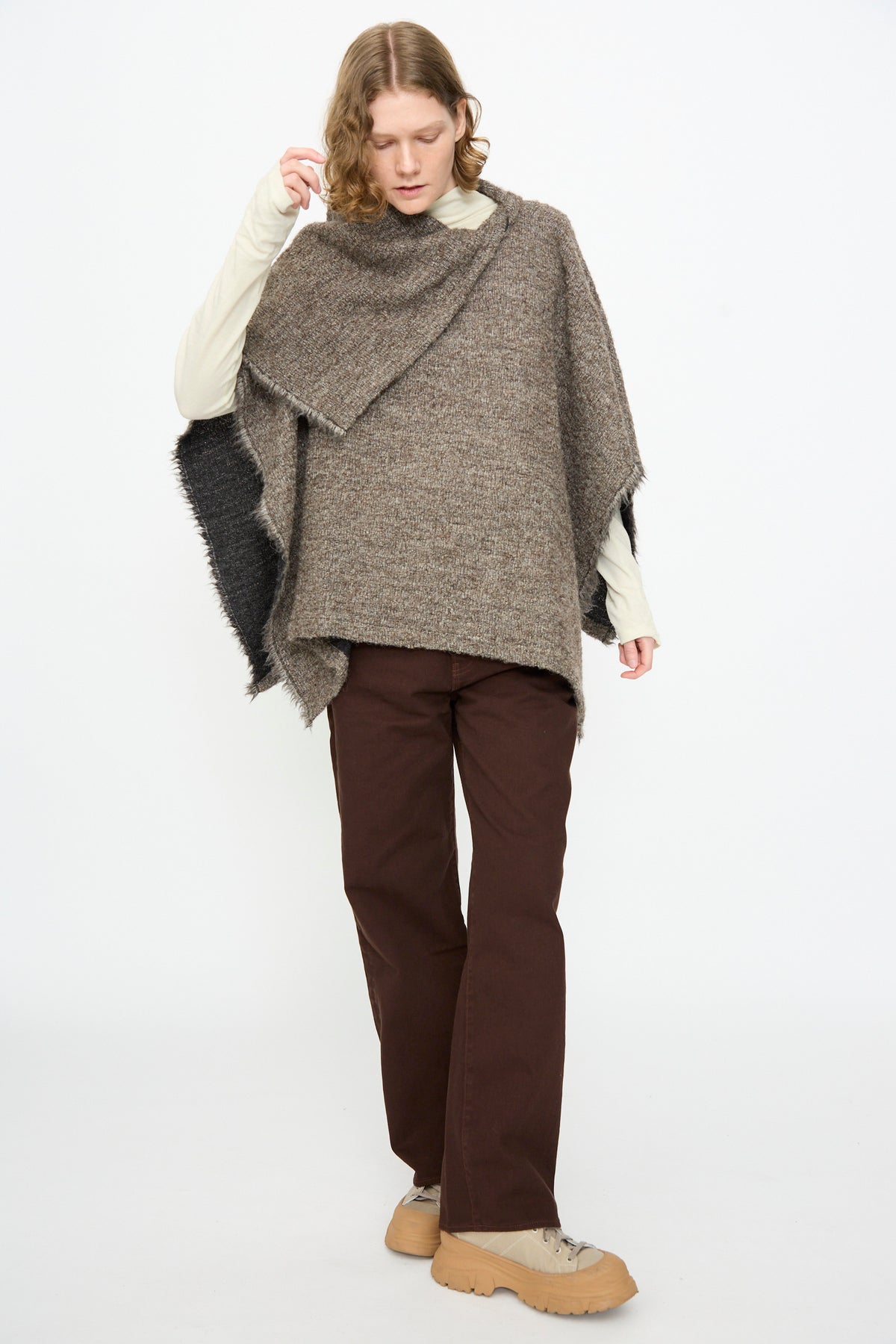 A person wearing a beige turtleneck, Black Crane Alpaca Short Poncho in Beige, brown pants, and tan shoes stands against a plain white background.