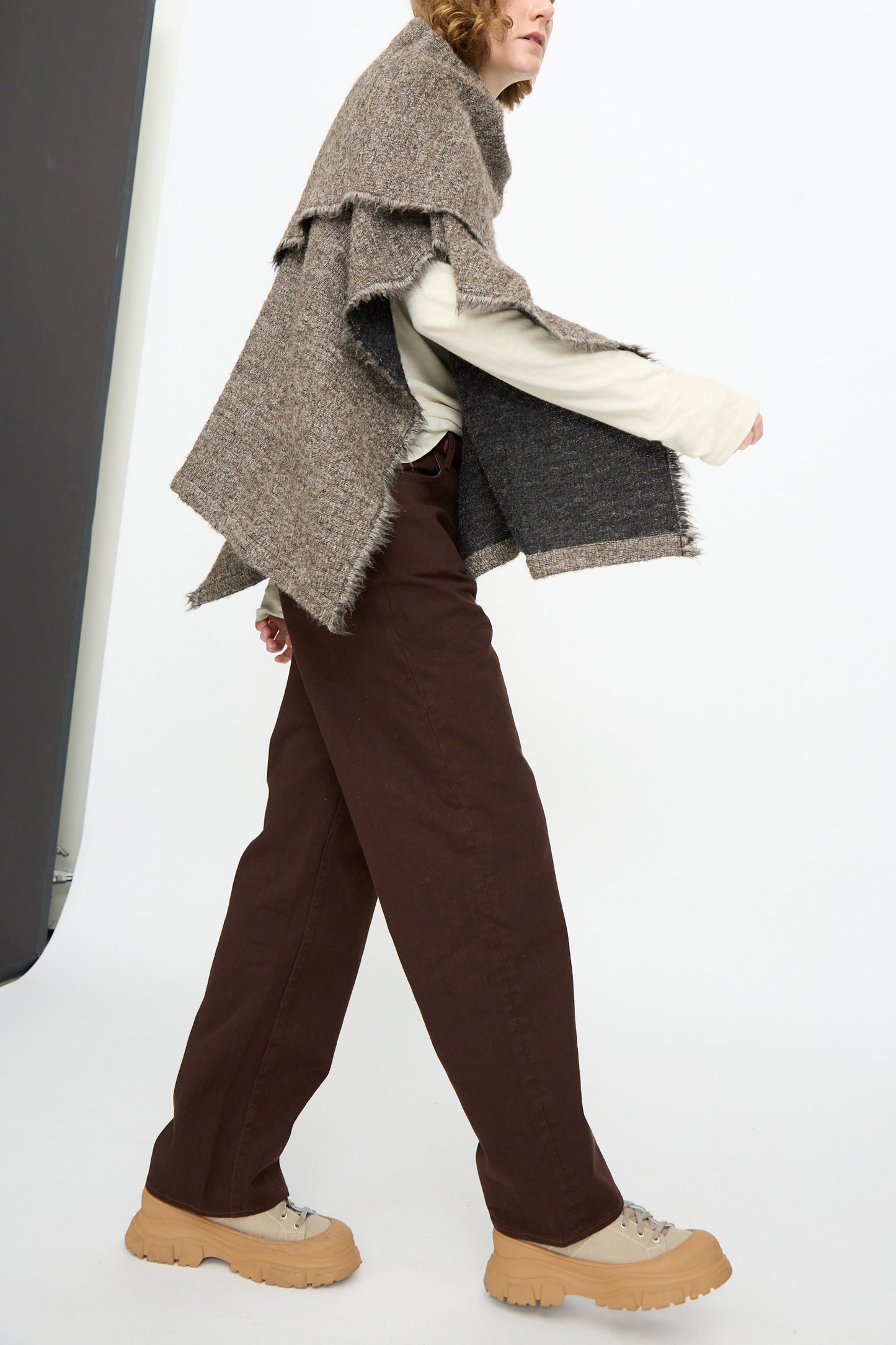 A person wears the Black Crane Alpaca Short Poncho in Beige layered over a beige sweater, paired with brown pants and beige platform shoes, while walking in a minimalist studio setting.
