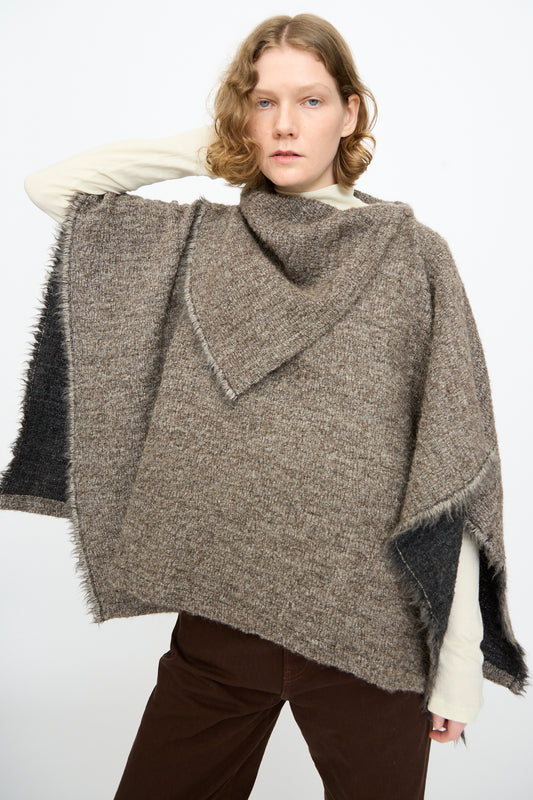 A person with short, wavy hair wears the Black Crane Alpaca Short Poncho in Beige over a cream long-sleeve top and brown pants, standing against a plain white background.