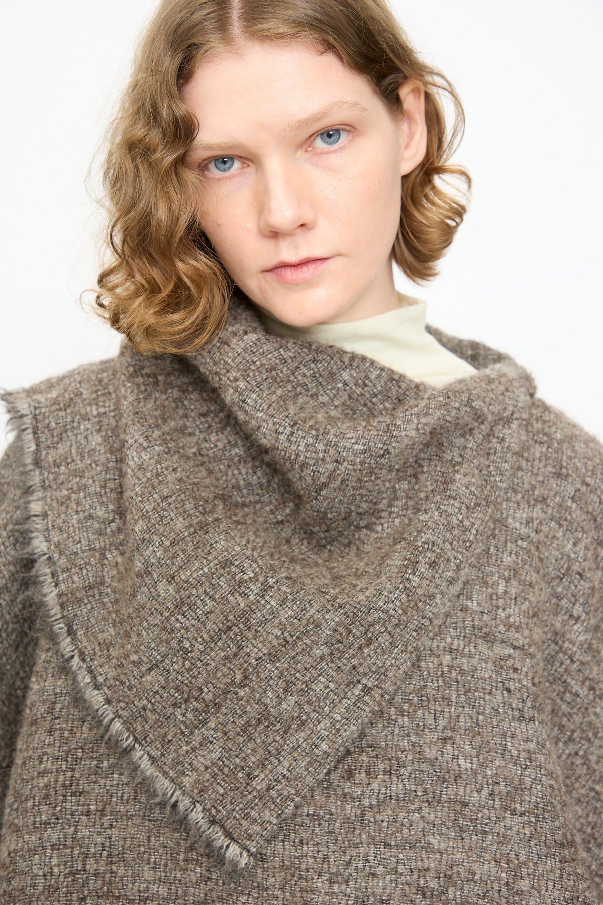 A person with short, wavy light brown hair wears the Black Crane Alpaca Short Poncho in Beige, featuring textured alpaca wool and fringe detailing, layered over a light turtleneck while looking at the camera against a plain background.