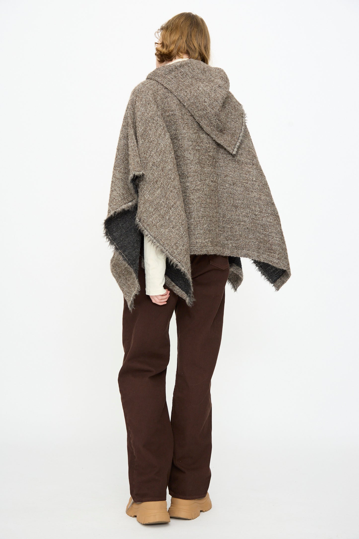 A person is seen from behind, wearing the Black Crane Alpaca Short Poncho in Beige—a textured alpaca wool poncho with a hood—paired with a cream long-sleeve shirt, dark brown pants, and tan shoes, all set against a plain white background. This cozy fringed poncho is made in the USA.