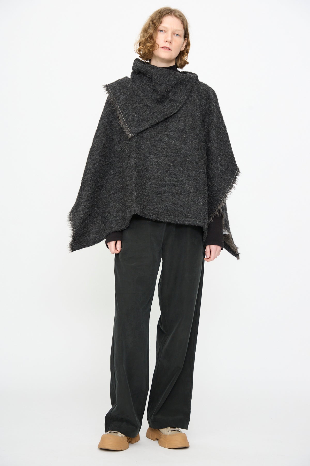 A person stands against a white background wearing the Black Crane Alpaca Short Poncho in Dark Grey, loose black pants, and tan shoes.