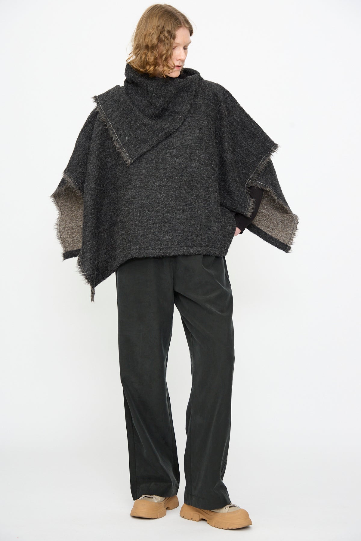 A person wearing the Black Crane Alpaca Short Poncho in Dark Grey, paired with wide-leg dark pants and tan shoes, stands against a plain white background.