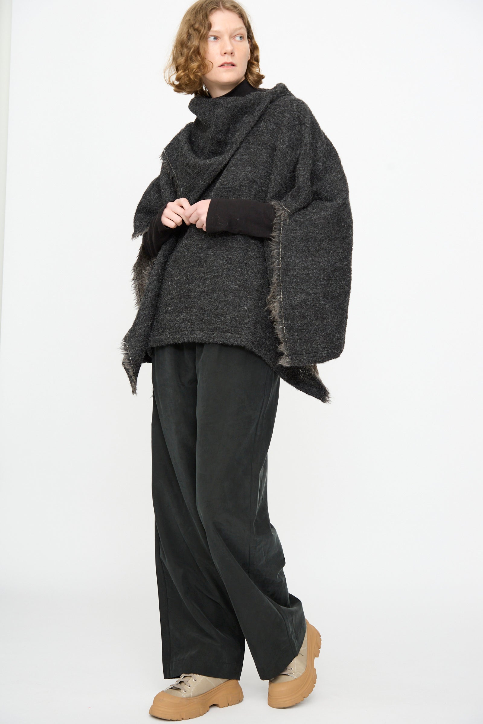 A person stands against a white background wearing the Black Crane Alpaca Short Poncho in Dark Grey, wide black pants, and beige shoes.