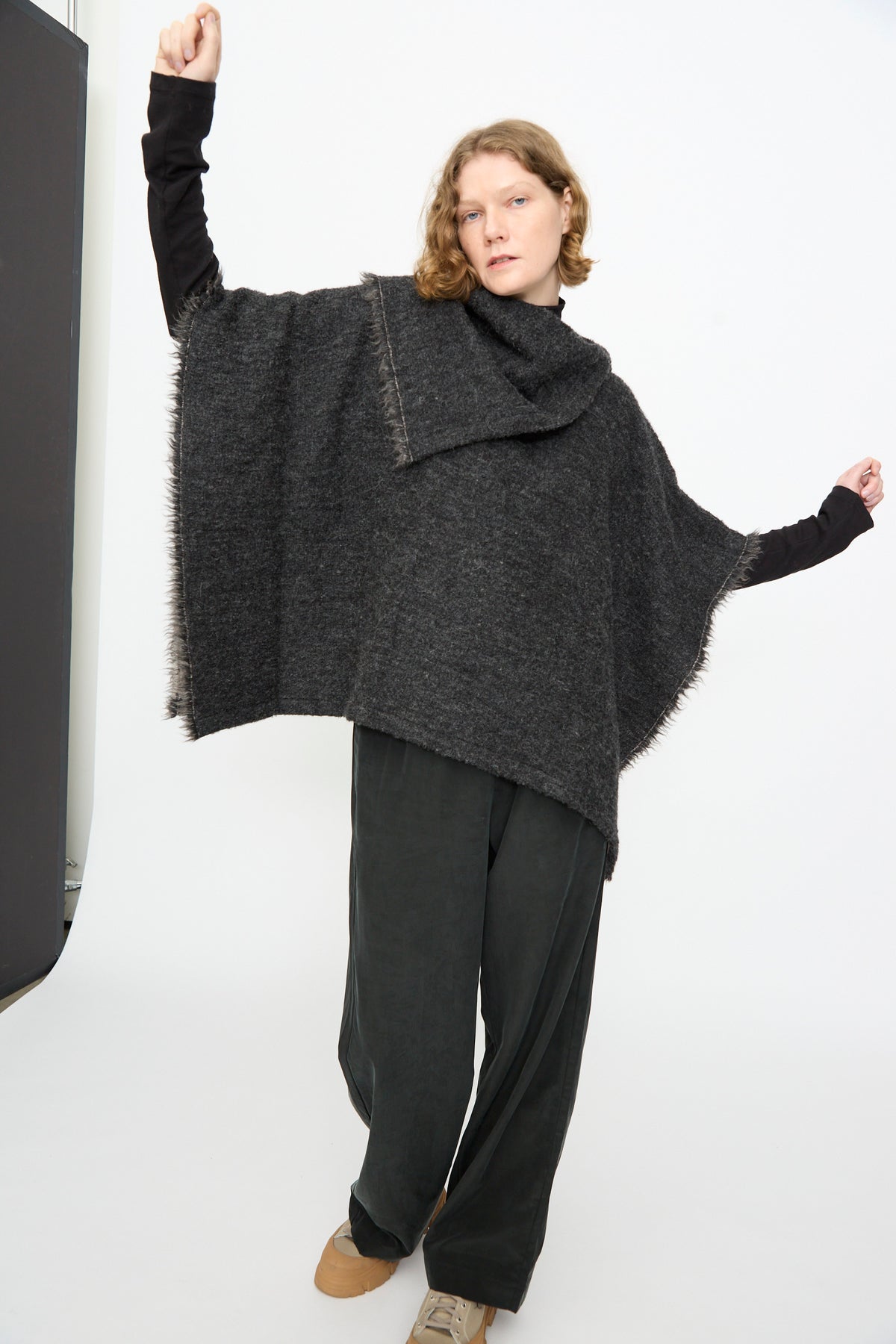 A person stands with arms raised against a plain white background, wearing the Black Crane Alpaca Short Poncho in Dark Grey over a black long-sleeve shirt, paired with wide-leg dark pants and beige shoes.