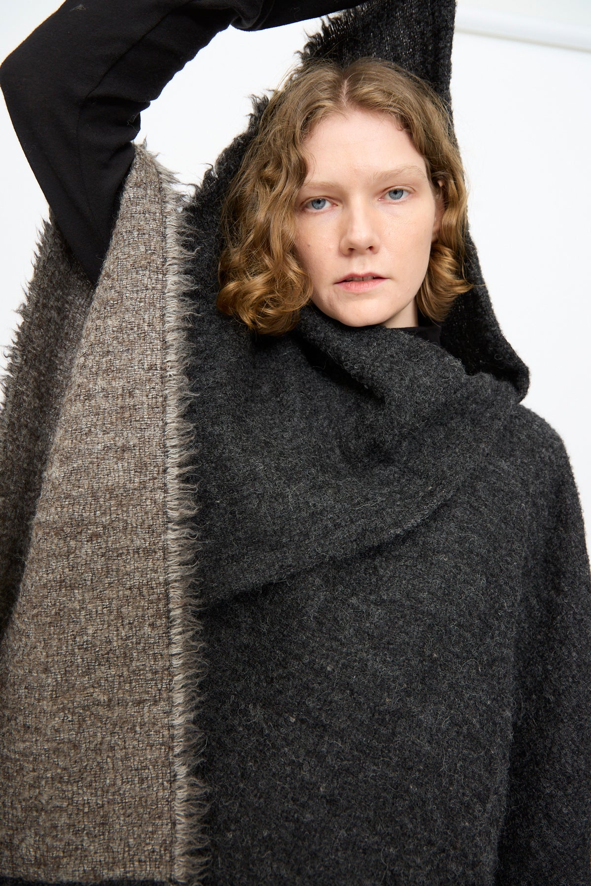 A person with wavy hair wears the Black Crane Alpaca Short Poncho in Dark Grey—featuring a textured, dark grey and brown alpaca wool design—raising one arm above their head and looking directly at the camera against a plain background.