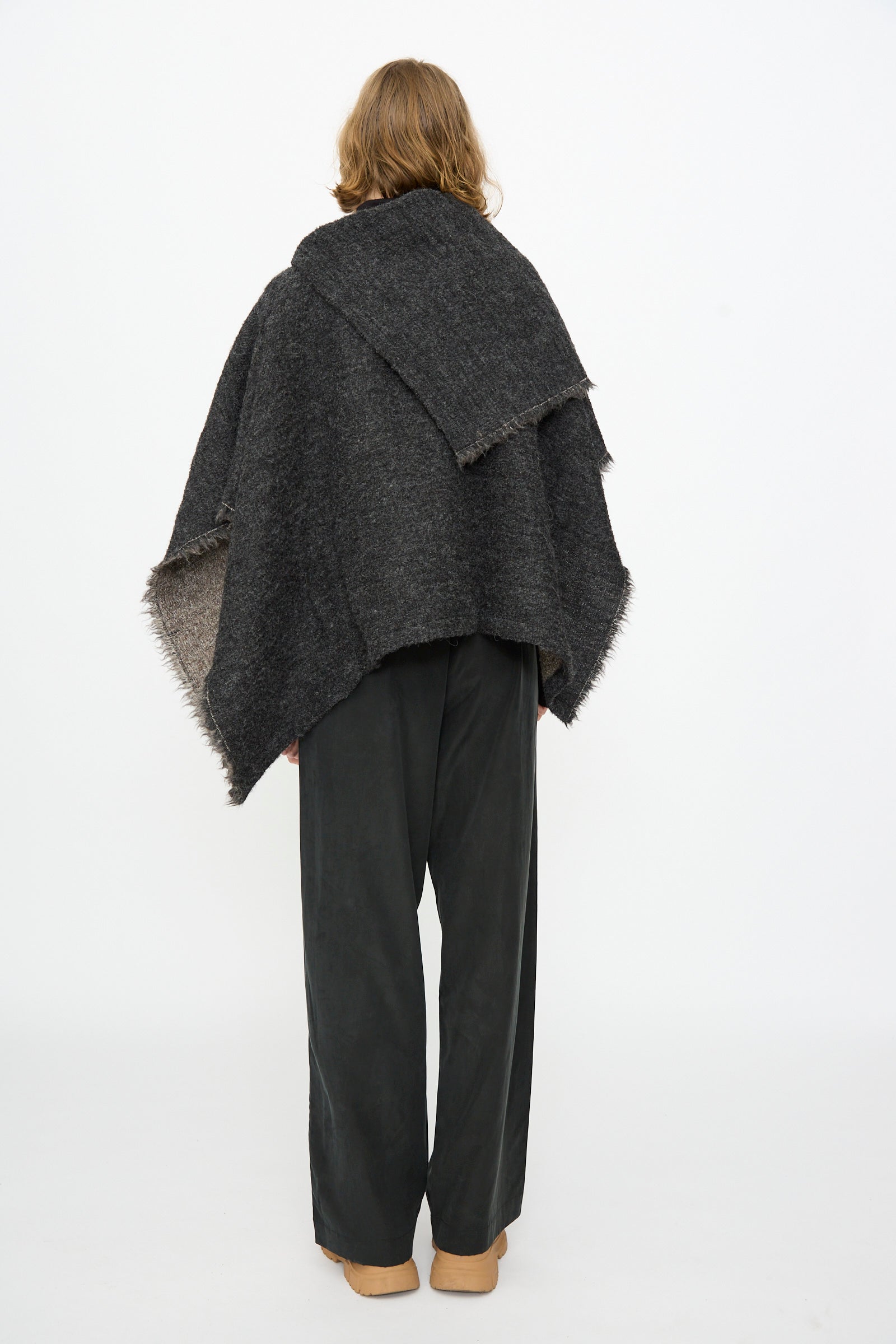 A person with shoulder-length hair stands facing away against a plain white background, wearing the Black Crane Alpaca Short Poncho in Dark Grey paired with loose black pants and tan shoes.