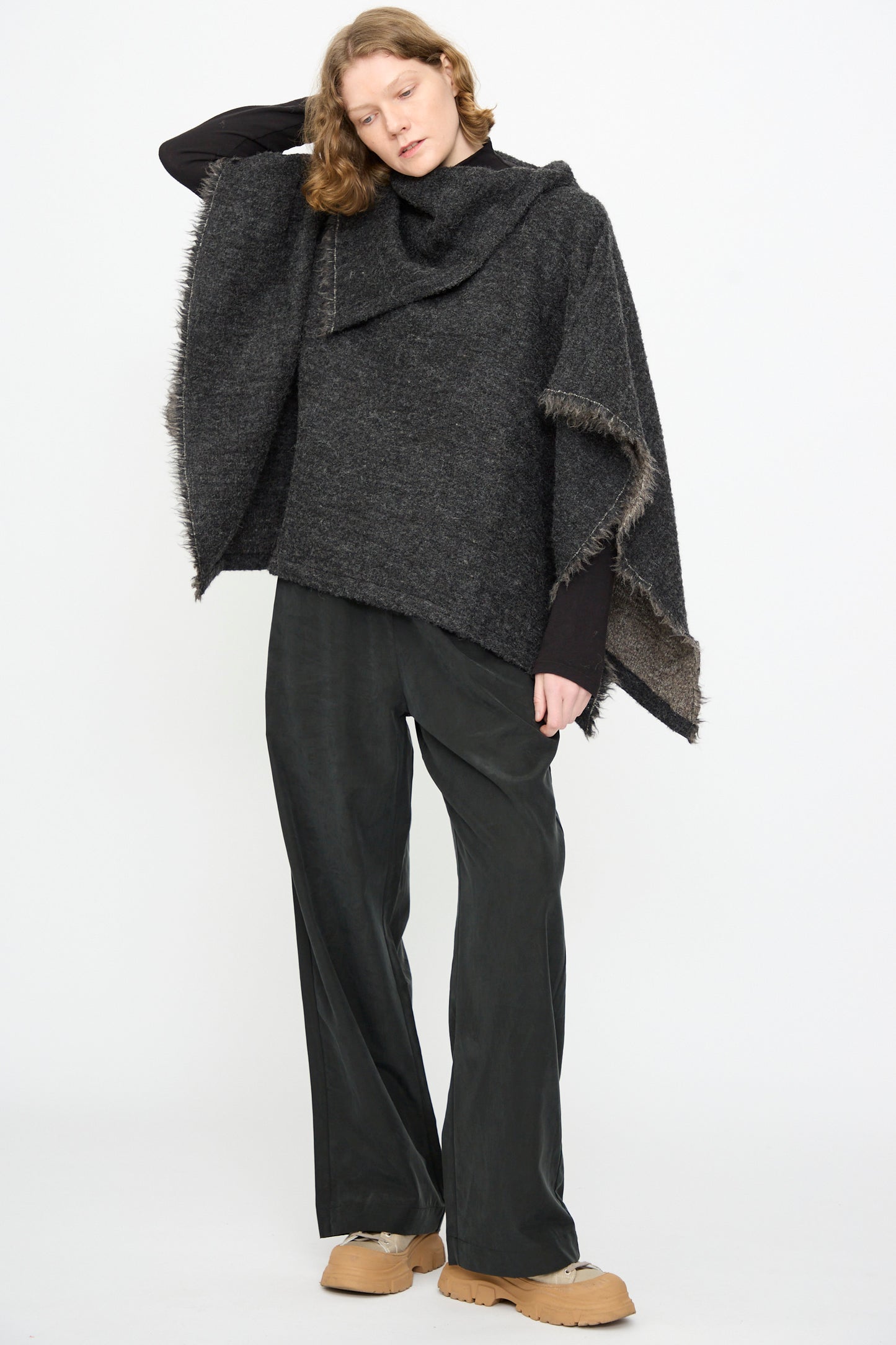 A person wearing the Black Crane Alpaca Short Poncho in Dark Grey, black wide-leg pants, and tan shoes stands against a plain white background.