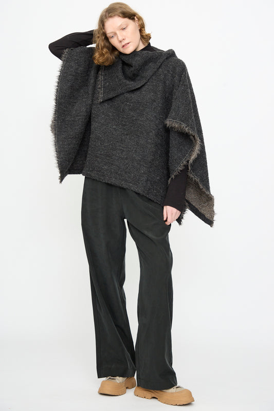 A person wearing the Black Crane Alpaca Short Poncho in Dark Grey, black wide-leg pants, and tan shoes stands against a plain white background.