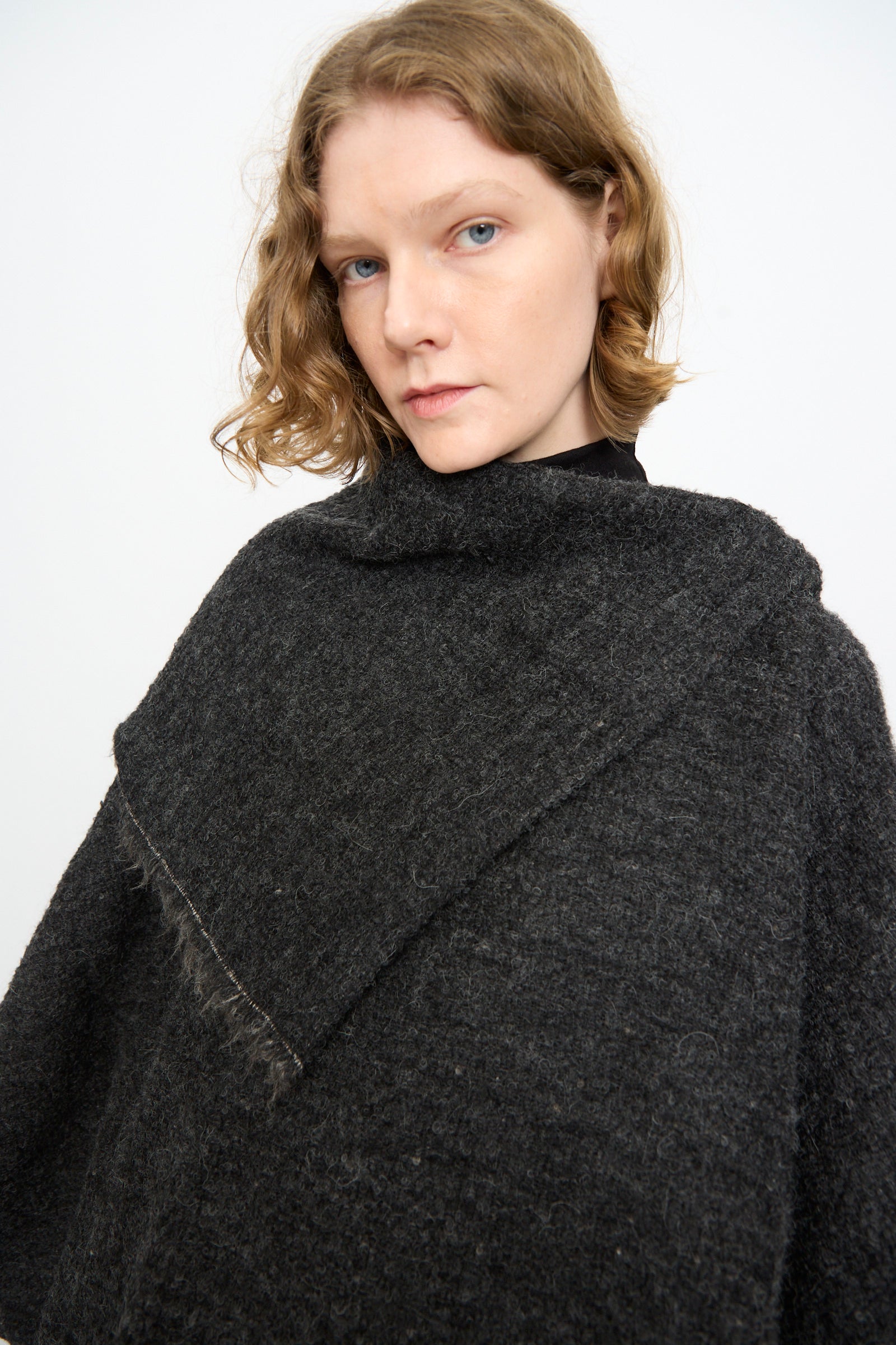A person with short, wavy, light brown hair models the Black Crane Alpaca Short Poncho in Dark Grey over a black top, standing against a plain white background.