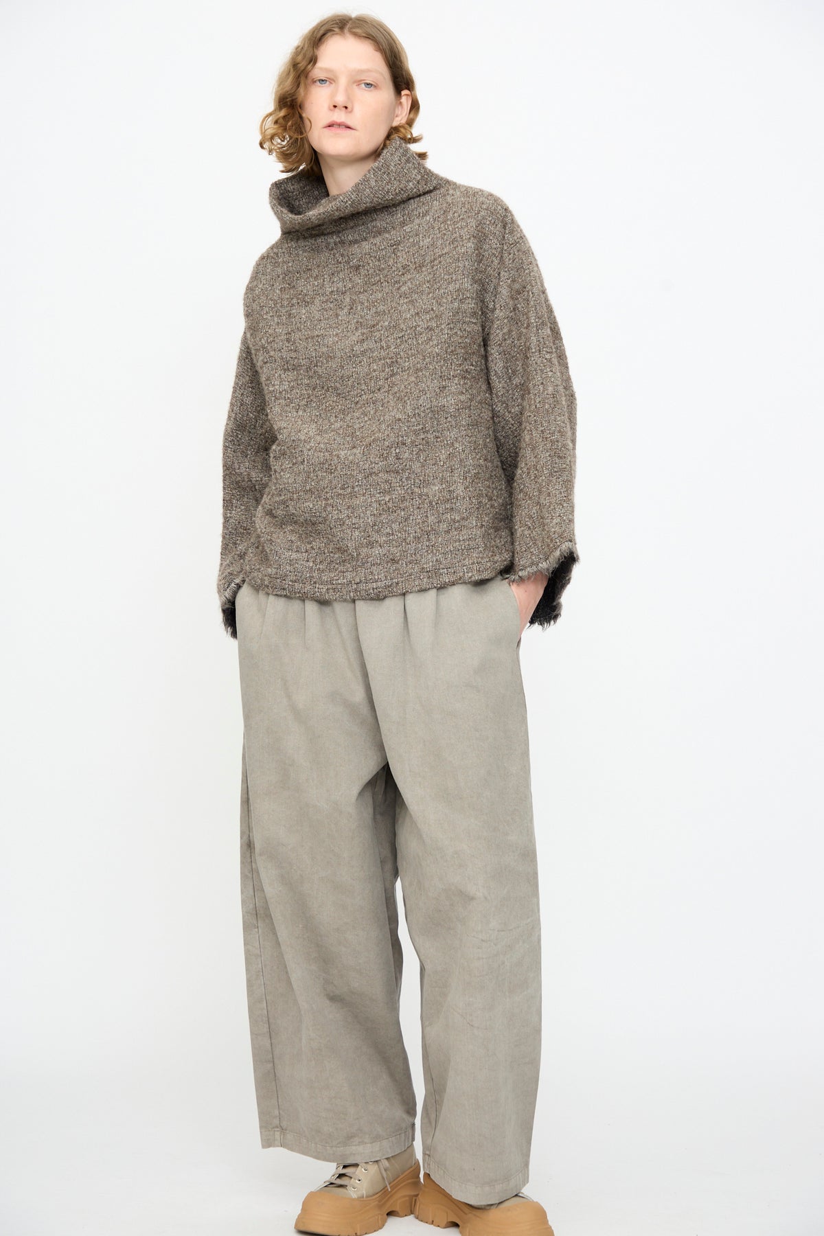 Person standing against a white background, wearing the Black Crane Alpaca Tube Top in Beige—a loose alpaca wool turtleneck tube top with a stylish fringed hem—paired with wide beige pants and tan shoes.