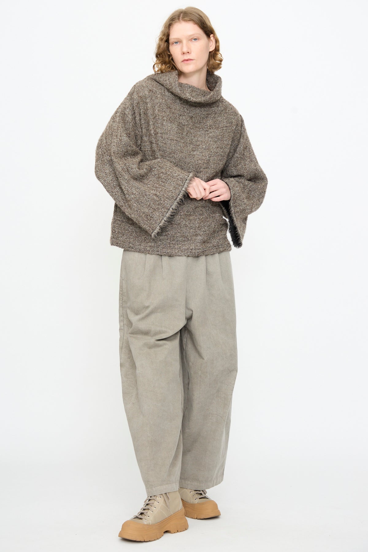 A person stands against a plain background, wearing the Black Crane Alpaca Tube Top in Beige, paired with wide gray pants and tan lace-up shoes.