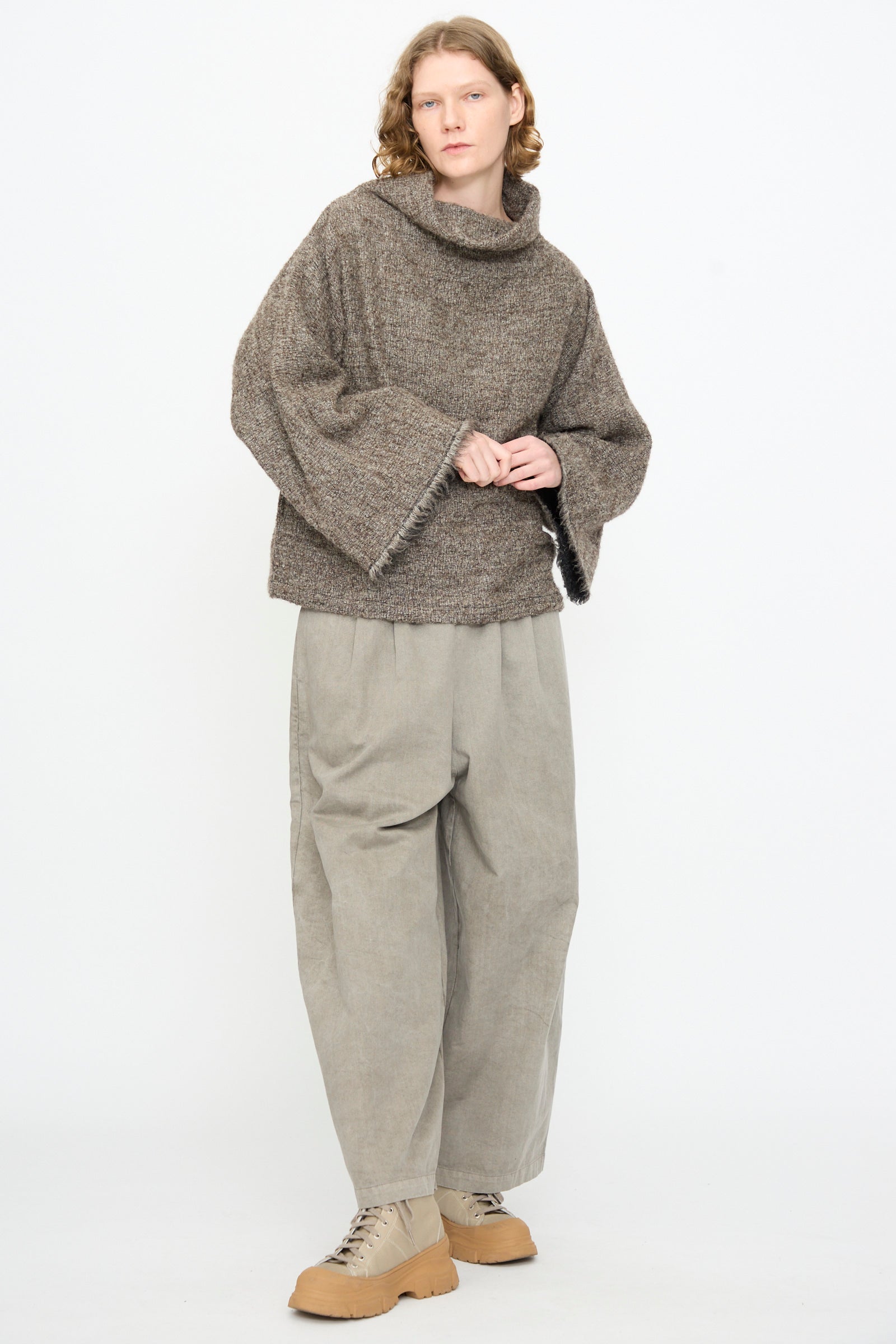A person stands against a plain background, wearing the Black Crane Alpaca Tube Top in Beige, paired with wide gray pants and tan lace-up shoes.