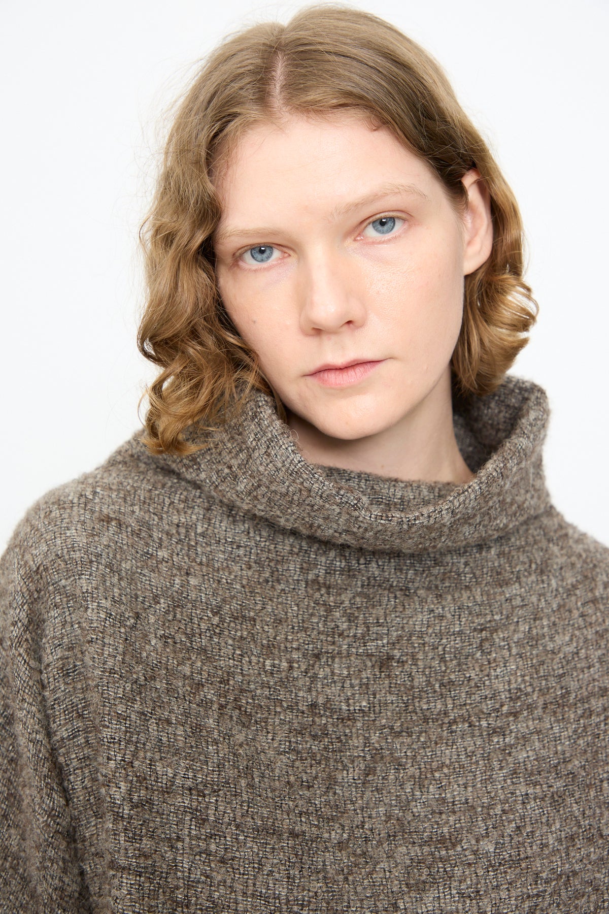 A person with short wavy light brown hair wears the Black Crane Alpaca Tube Top in Beige, featuring a textured knit and fringed hem, while looking at the camera against a plain white background.