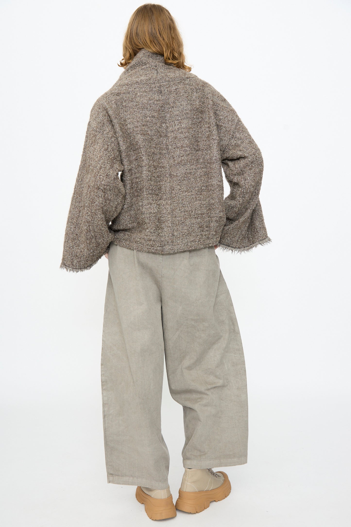 A person stands with their back to the camera, dressed in a loose brown jacket layered over the Black Crane Alpaca Tube Top in Beige, paired with wide-leg beige pants and tan shoes, set against a plain white background.
