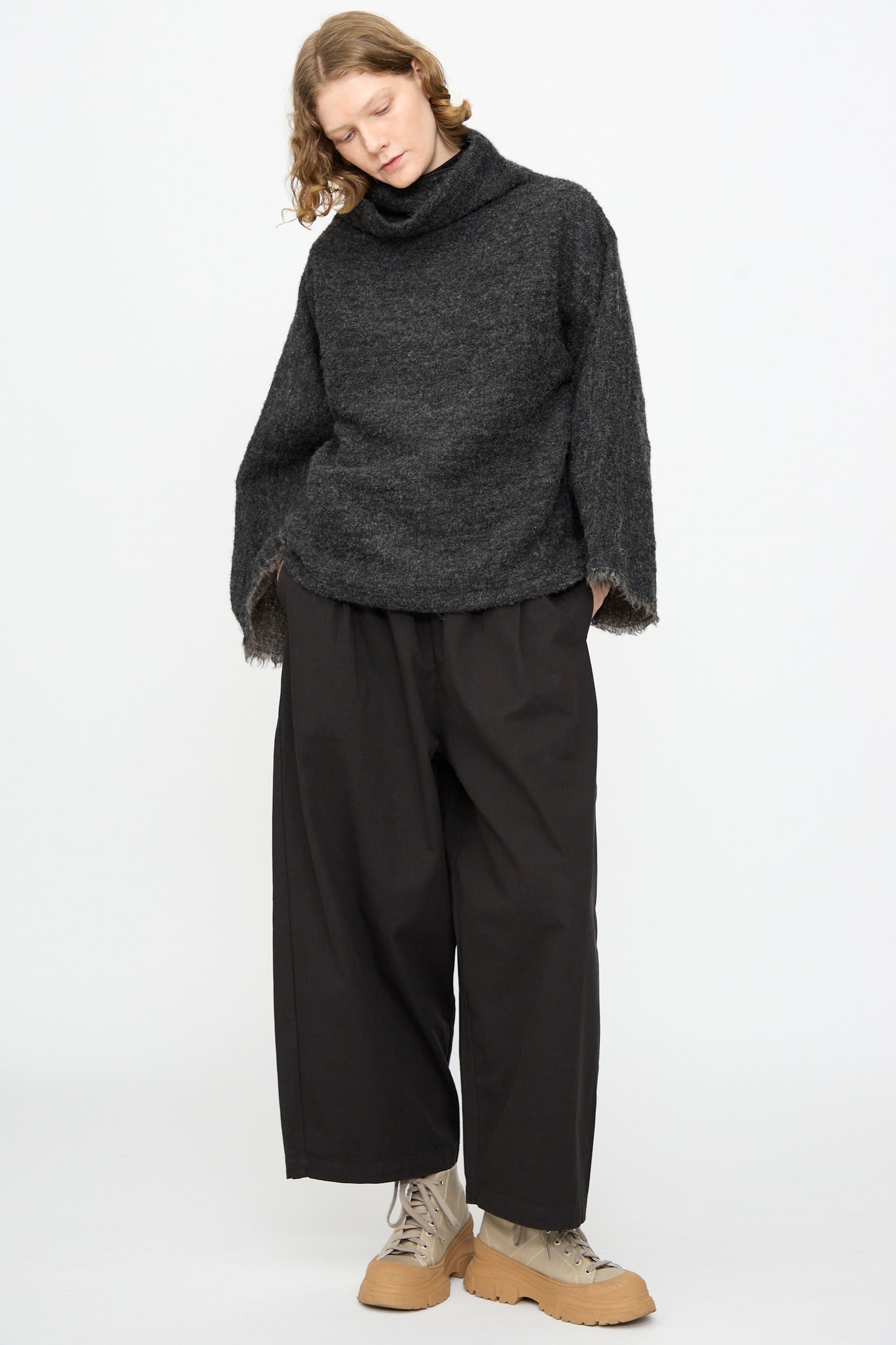 A person stands against a plain white background wearing the Black Crane Alpaca Tube Top in Dark Grey, paired with black wide-leg pants and light tan lace-up boots.