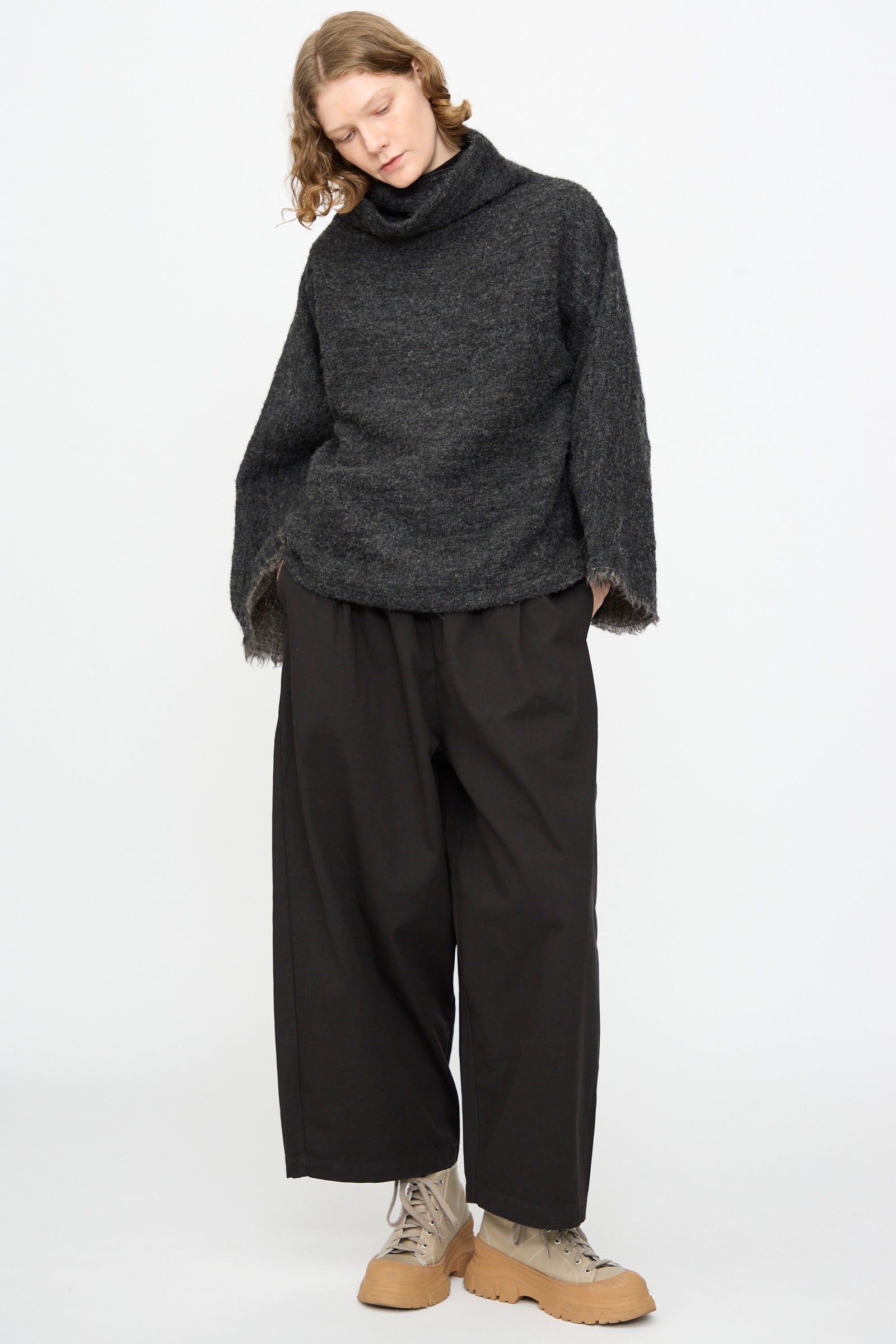 A person stands against a plain white background wearing the Black Crane Alpaca Tube Top in Dark Grey, paired with black wide-leg pants and light tan lace-up boots.
