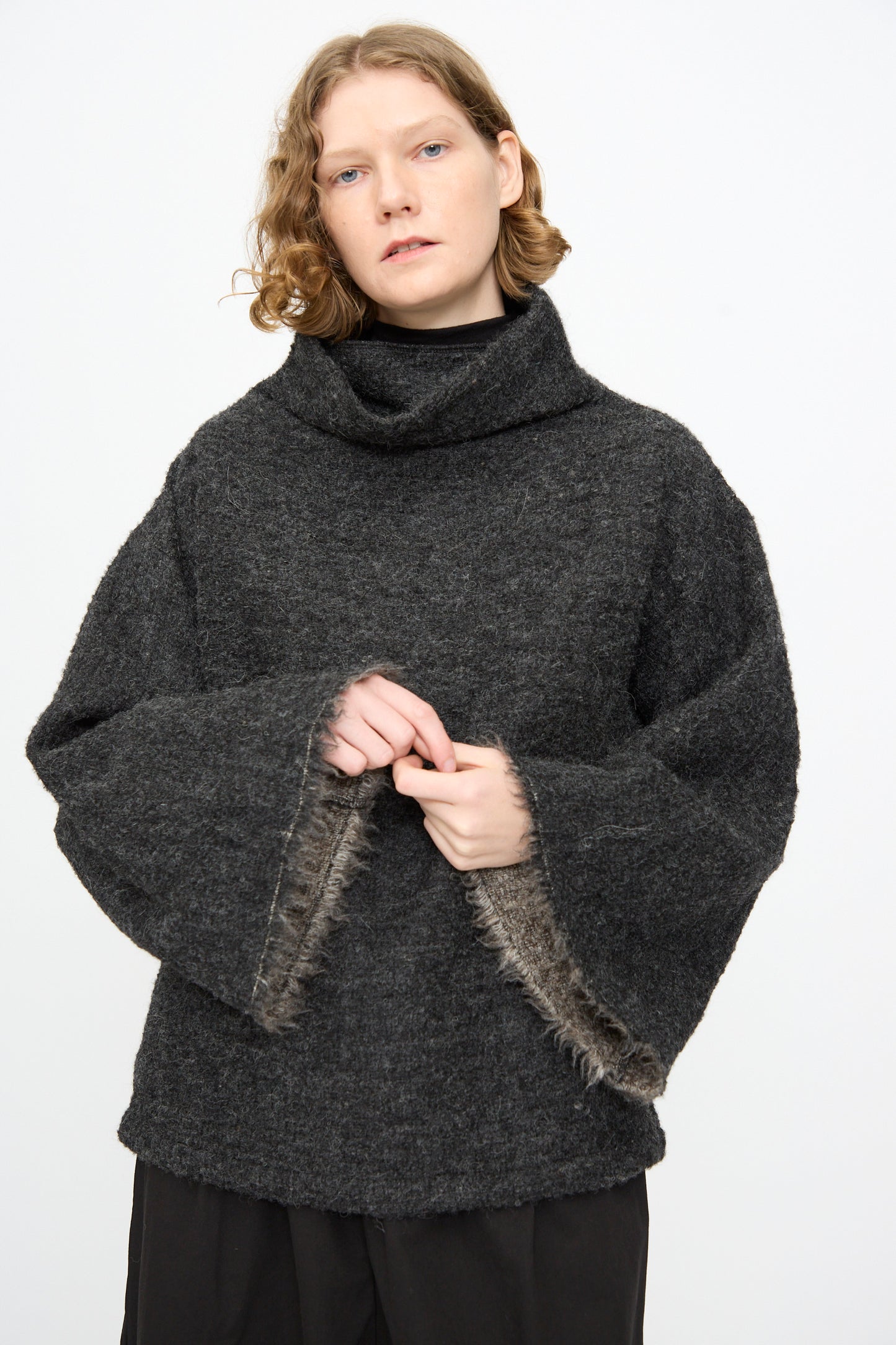 Wearing the Black Crane Alpaca Tube Top in Dark Grey, a person with short, wavy hair stands against a plain white background, layered under a loose sweater with wide sleeves and fur trim.